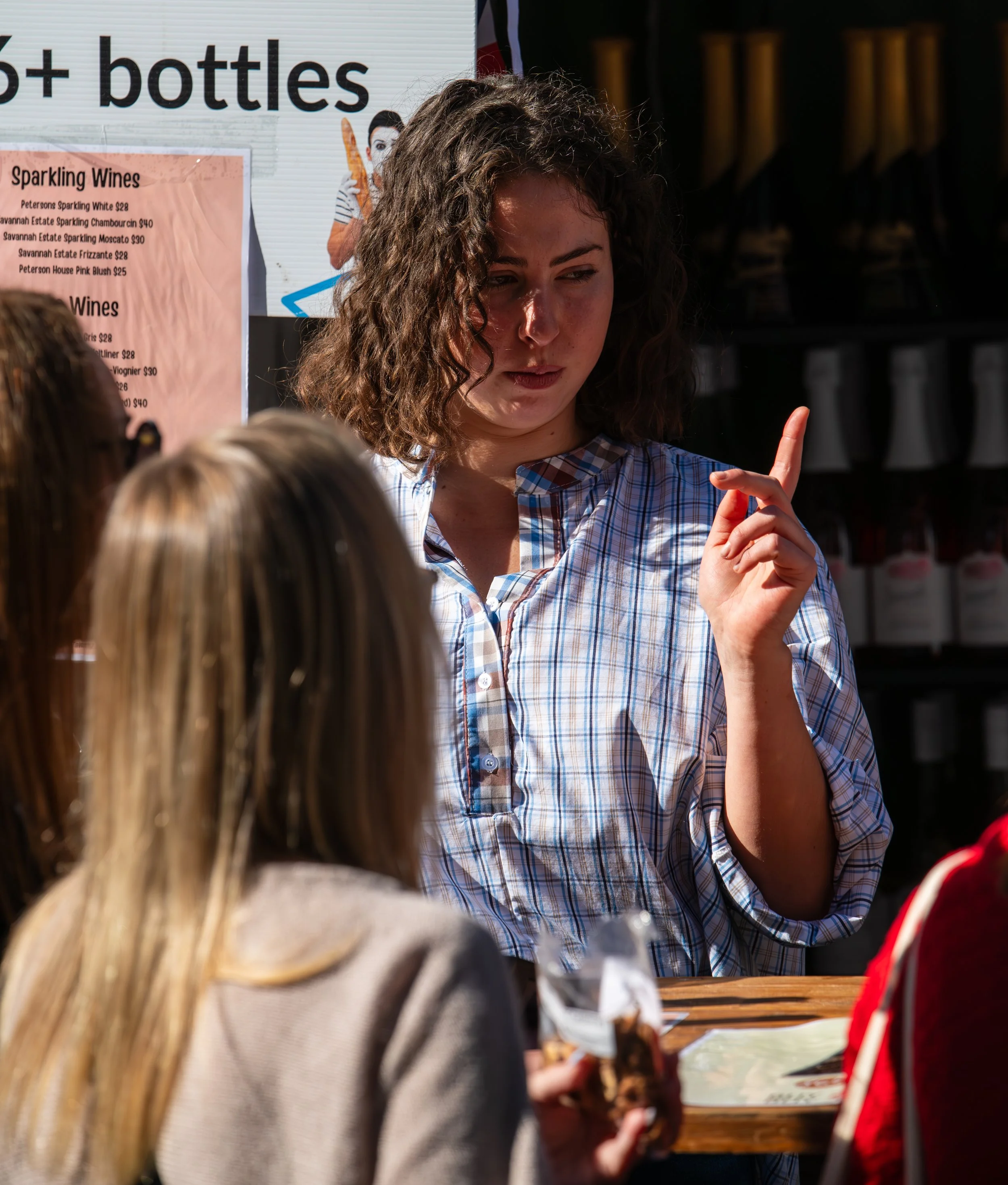 Young woman with curly hair wearing a blue and white plaid shirt, raising her index finger, talking to a group of girls at an outdoor event with a drink stand in the background.