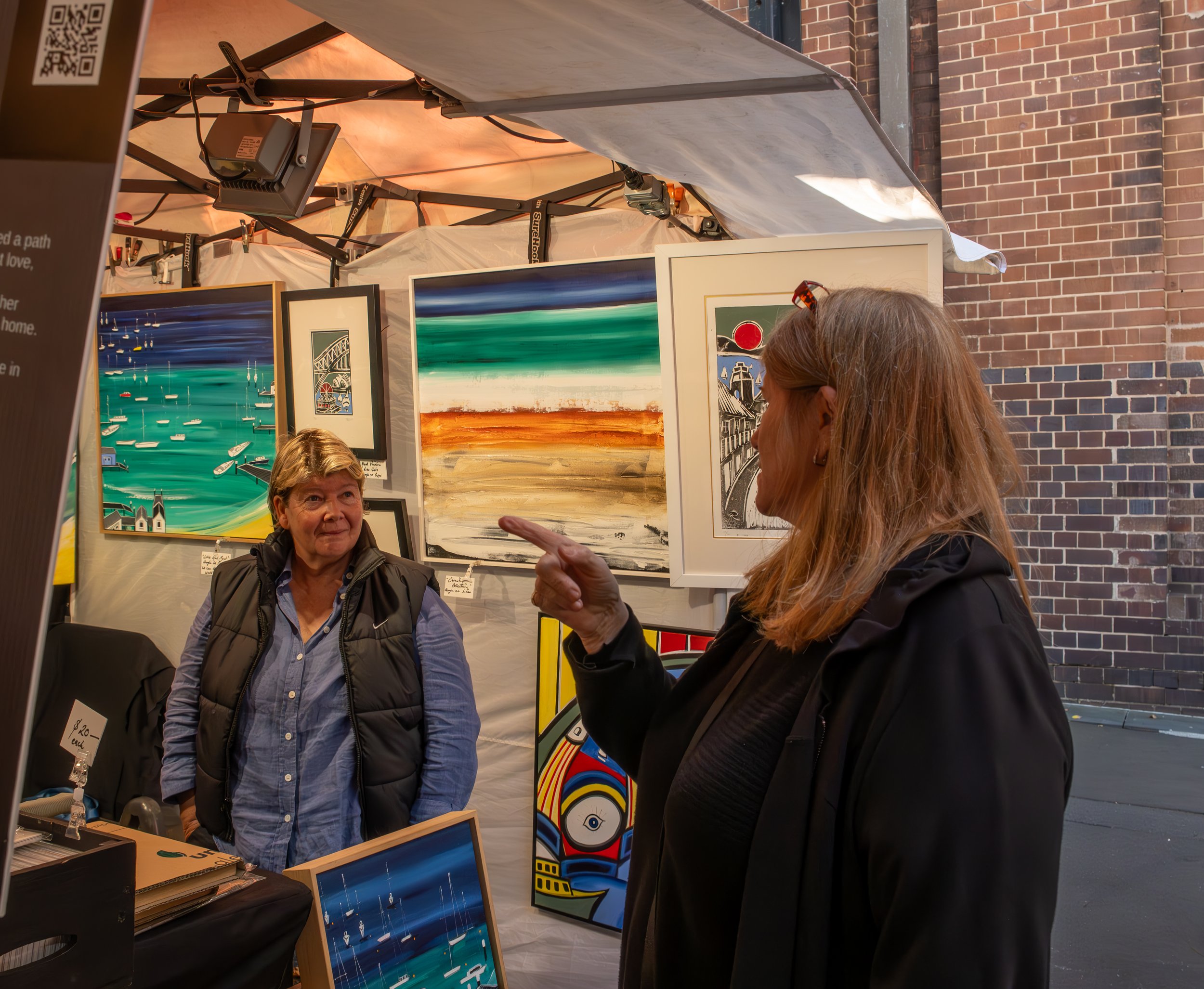 Two women conversing at an art booth featuring colorful paintings of boats, landscapes, and abstract designs, set against a brick wall background.
