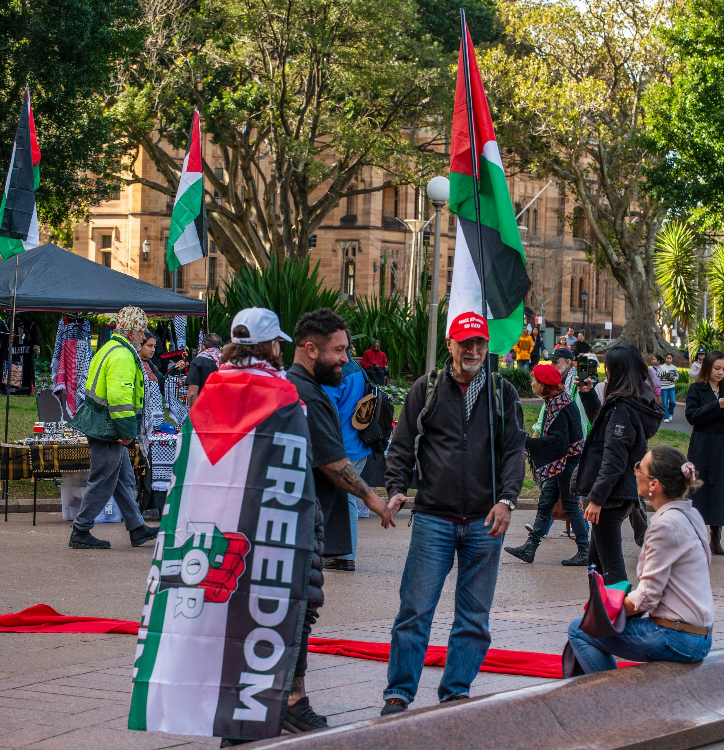 A group of people at a public gathering in a park, with some holding Palestinian flags and one person wearing a "Free Palestine" banner. There are trees, a historic building, and a vendor tent in the background.