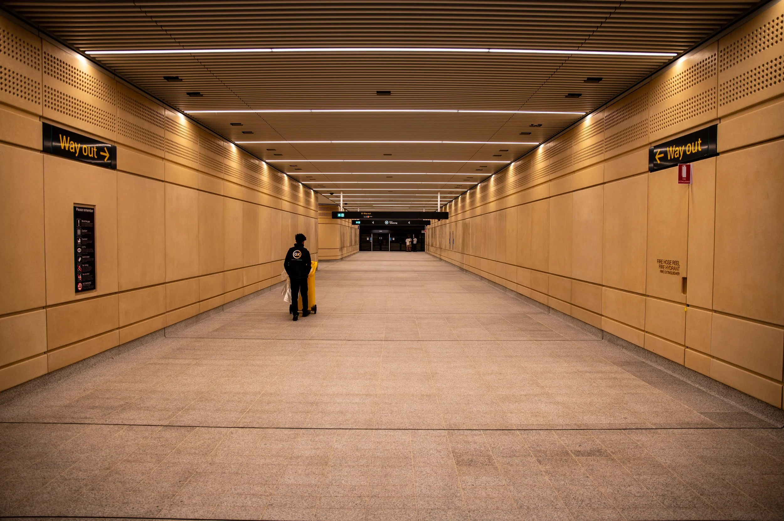 Empty underground subway station corridor with a person pushing a yellow cleaning cart and two other individuals in the distance. Signage indicating way out on both sides of the corridor. The ceiling has linear lighting, and the walls are beige with ventilation holes.
