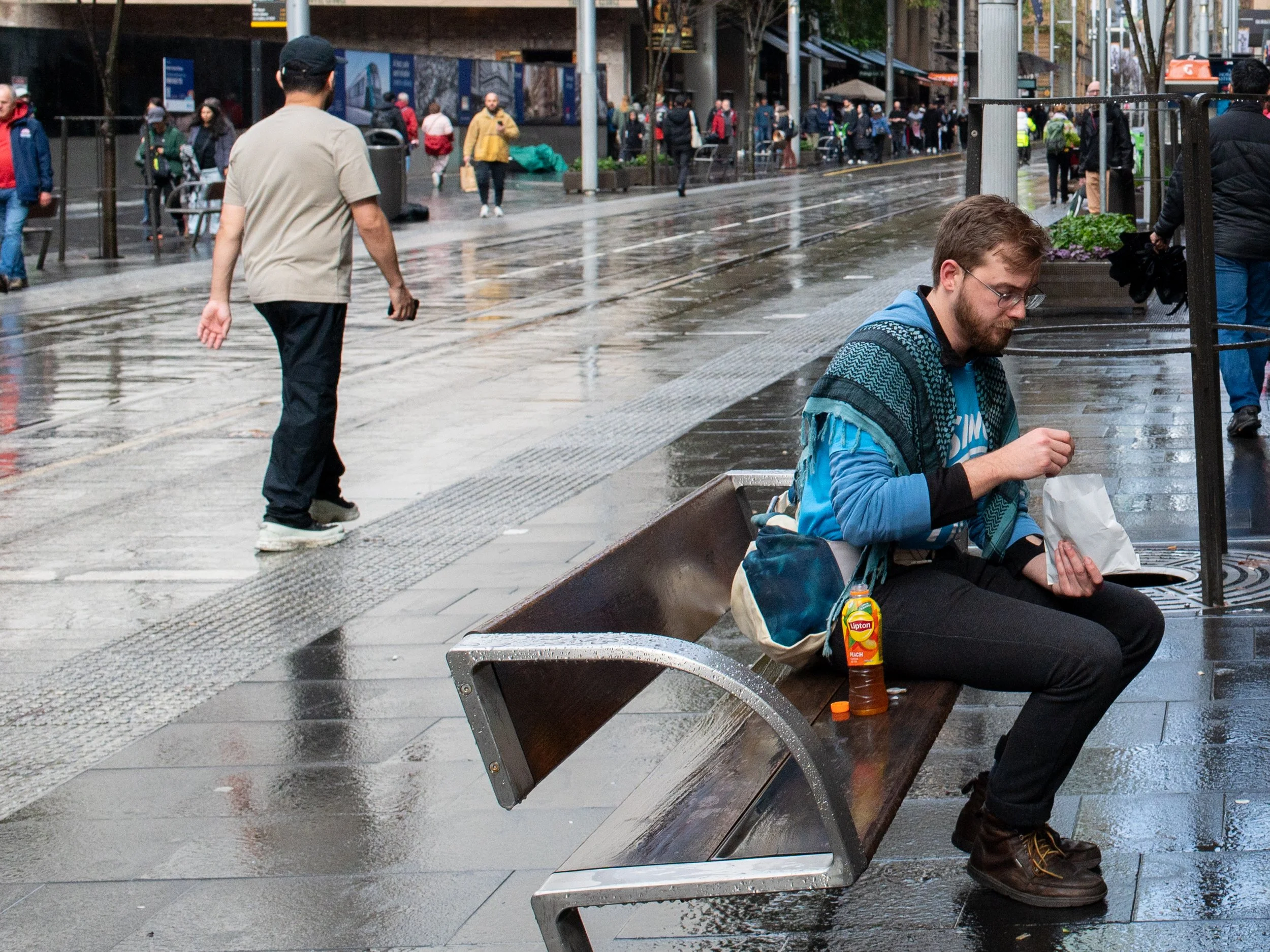 A man sitting on a rain-soaked park bench eating from a paper bag, beside a bottle of Lipton Iced Tea, in a busy city street with wet pavement and people walking in the background.