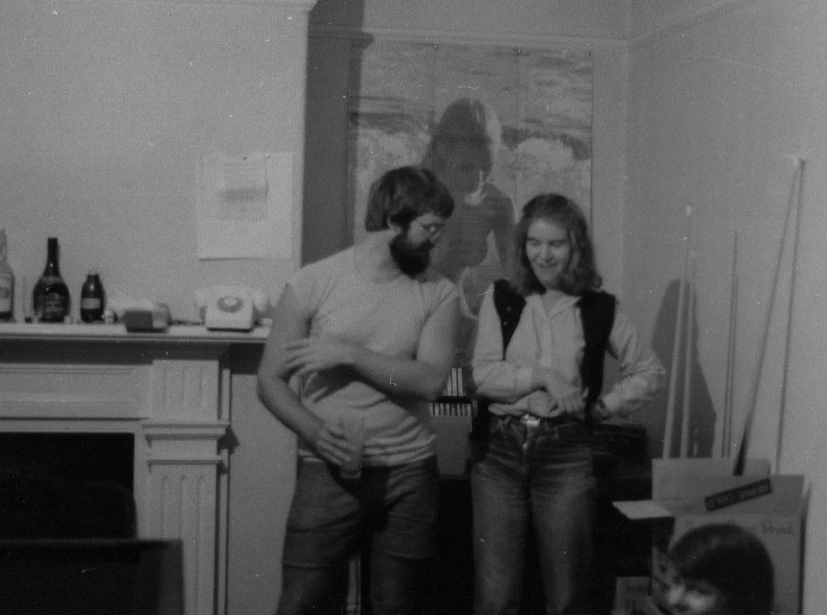 Two people, a man with glasses and a woman with a backpack, standing and talking in a room with a table and bottles in the background, while a third person, a child, is sitting on the floor nearby.