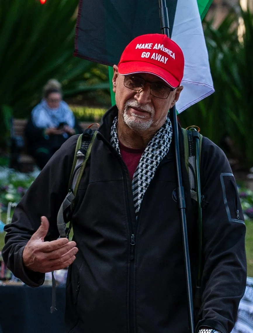 A man wearing a red 'Make America Go Away' hat, glasses, and a scarf, holding a flag and wearing a backpack. In the background, a woman is sitting on a bench outdoors.
