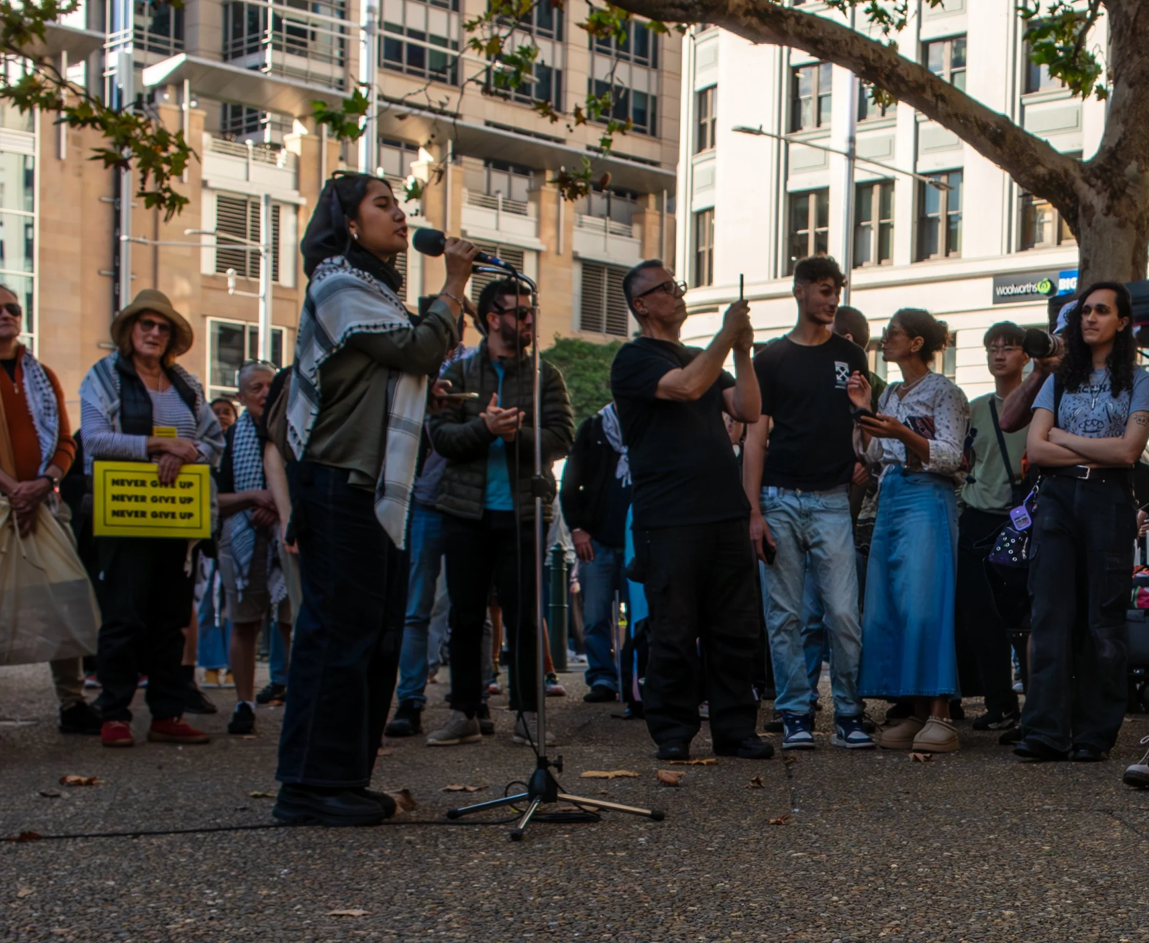 A diverse group of people gathered outdoors in an urban area, with some standing and one woman speaking into a microphone. The scene is set on a city street with trees and tall buildings in the background, indicating a protest or rally.
