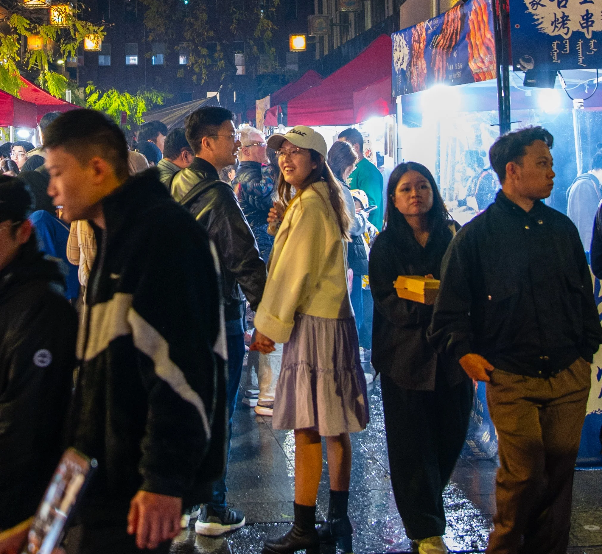 People walking through a lively night market with food stalls and red vendor tents on a city street, illuminated by bright lights.