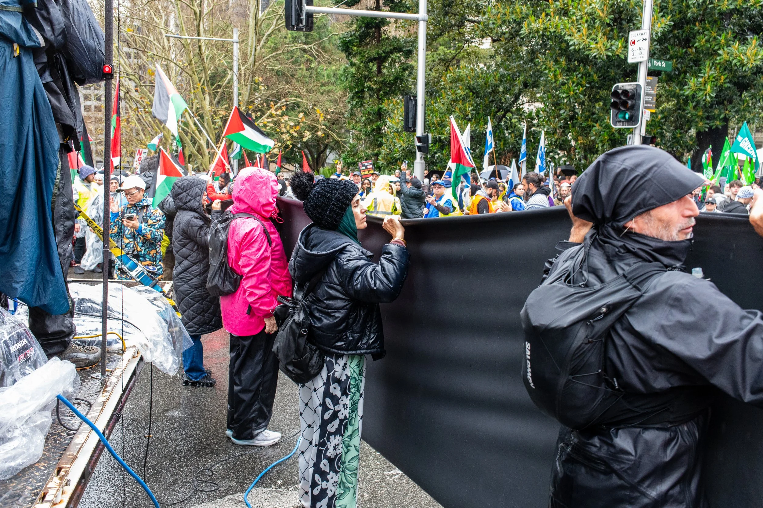 Protesters gather at a demonstration holding flags and a large black banner in a street with trees and traffic lights. Many are wearing rain jackets and hats.