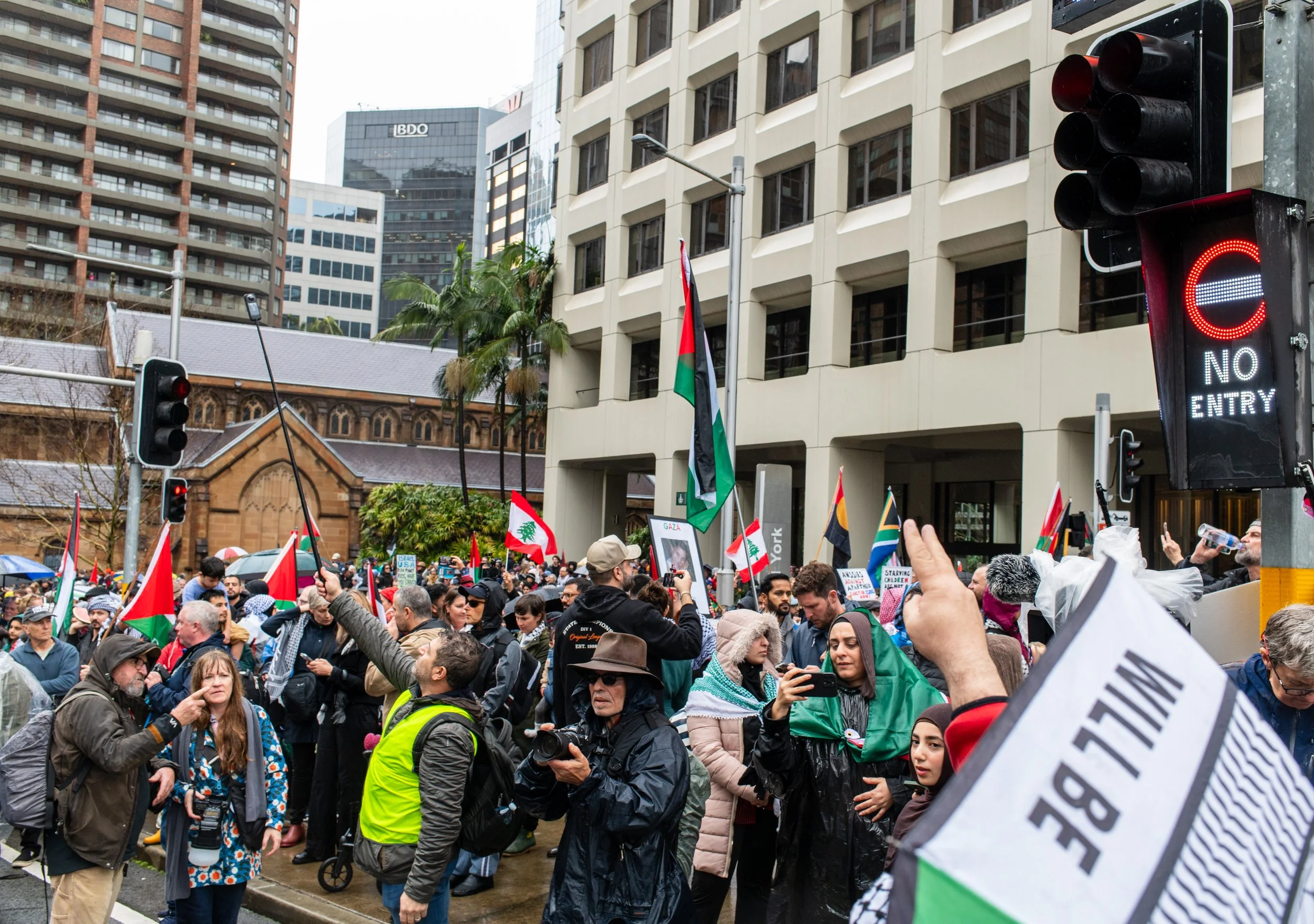 Crowd of protesters holding flags and signs in an urban area with tall buildings, some wearing raincoats and hats, and traffic lights displaying a no-entry sign.