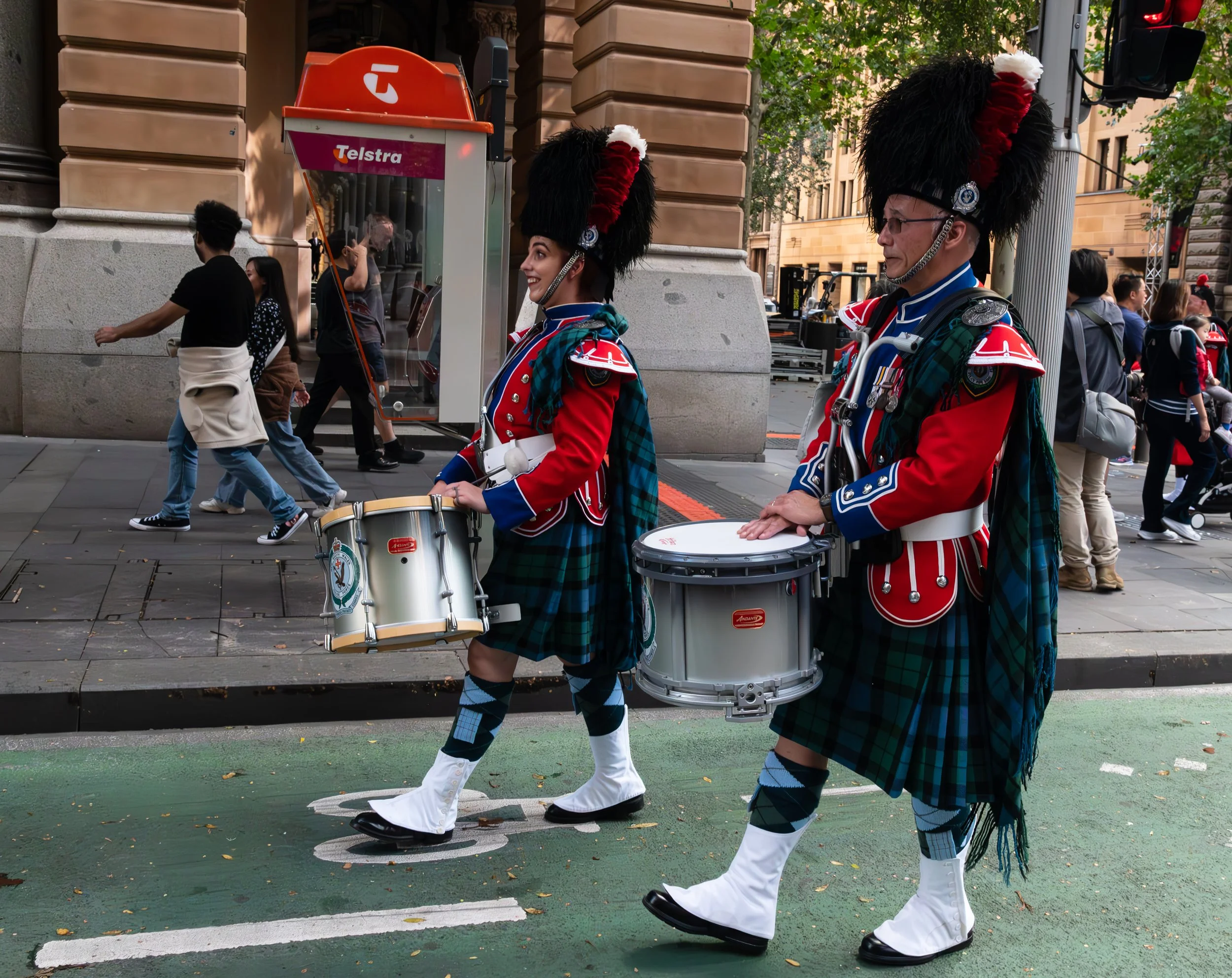 Two people dressed in traditional Scottish Highland dress, playing drums while walking in a parade on a city street.