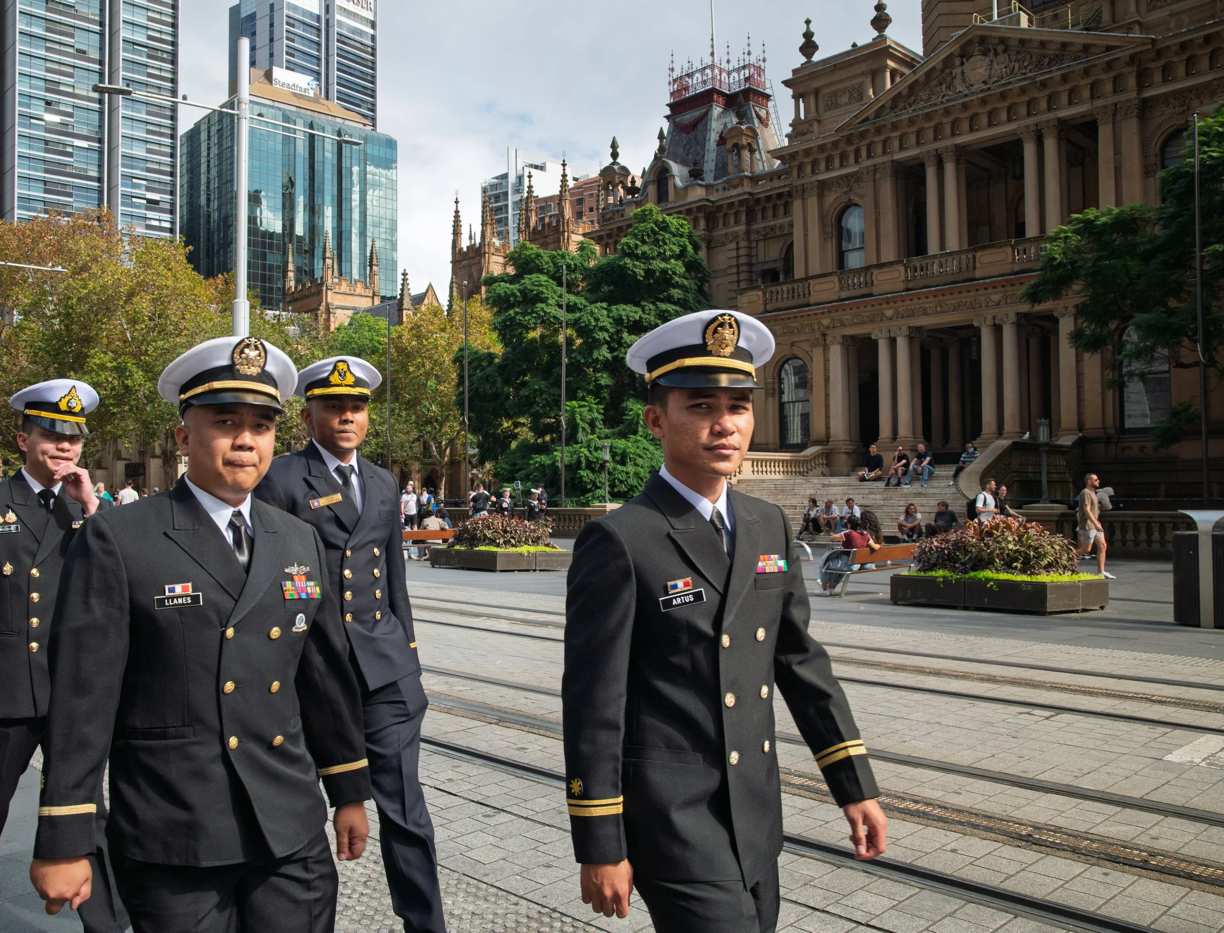A group of military personnel dressed in black uniforms and white hats walking outdoors in an urban setting with historic and modern buildings in the background.