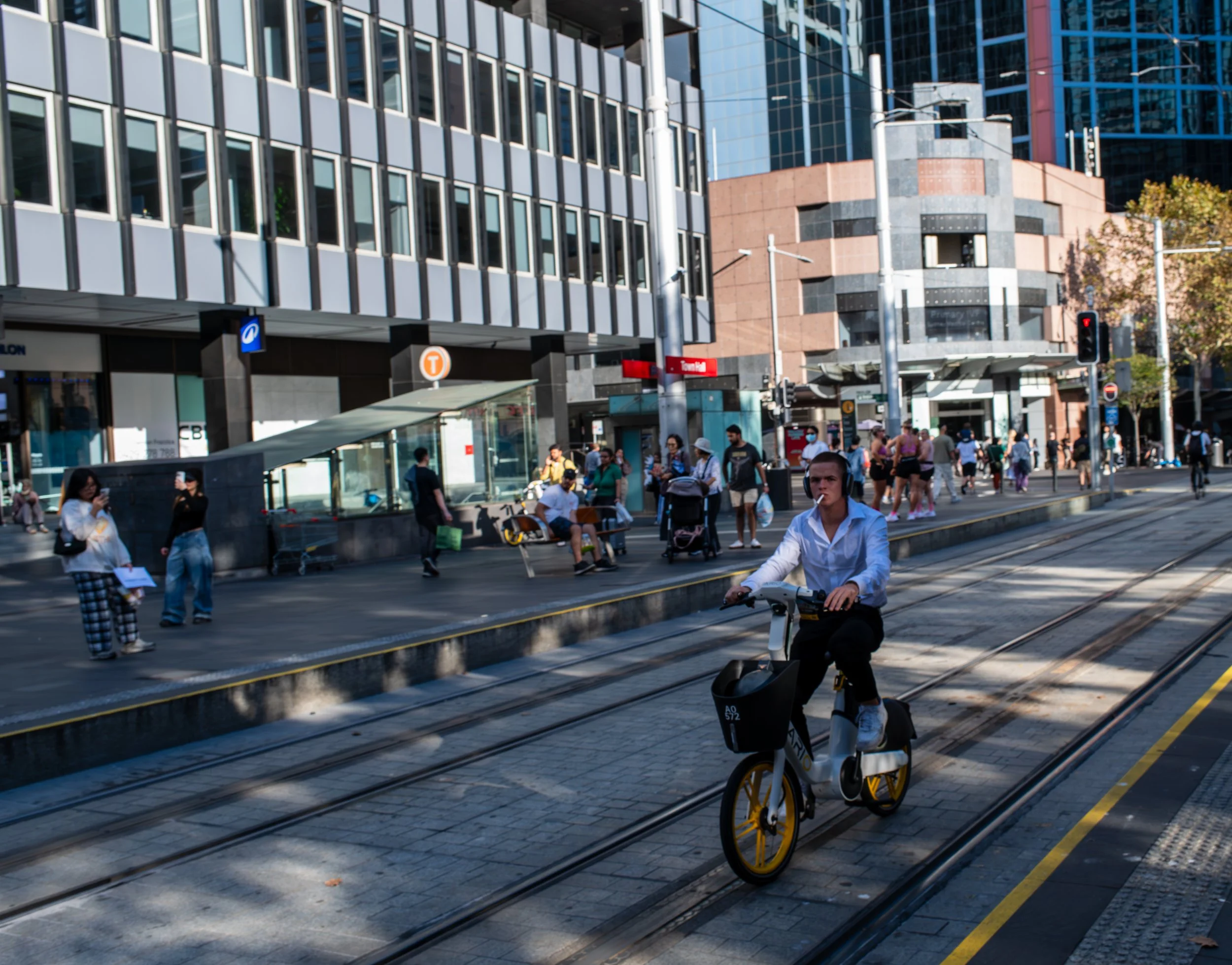 A busy city street with pedestrians waiting at a bus stop, a woman riding an electric bike, and tall modern buildings in the background.