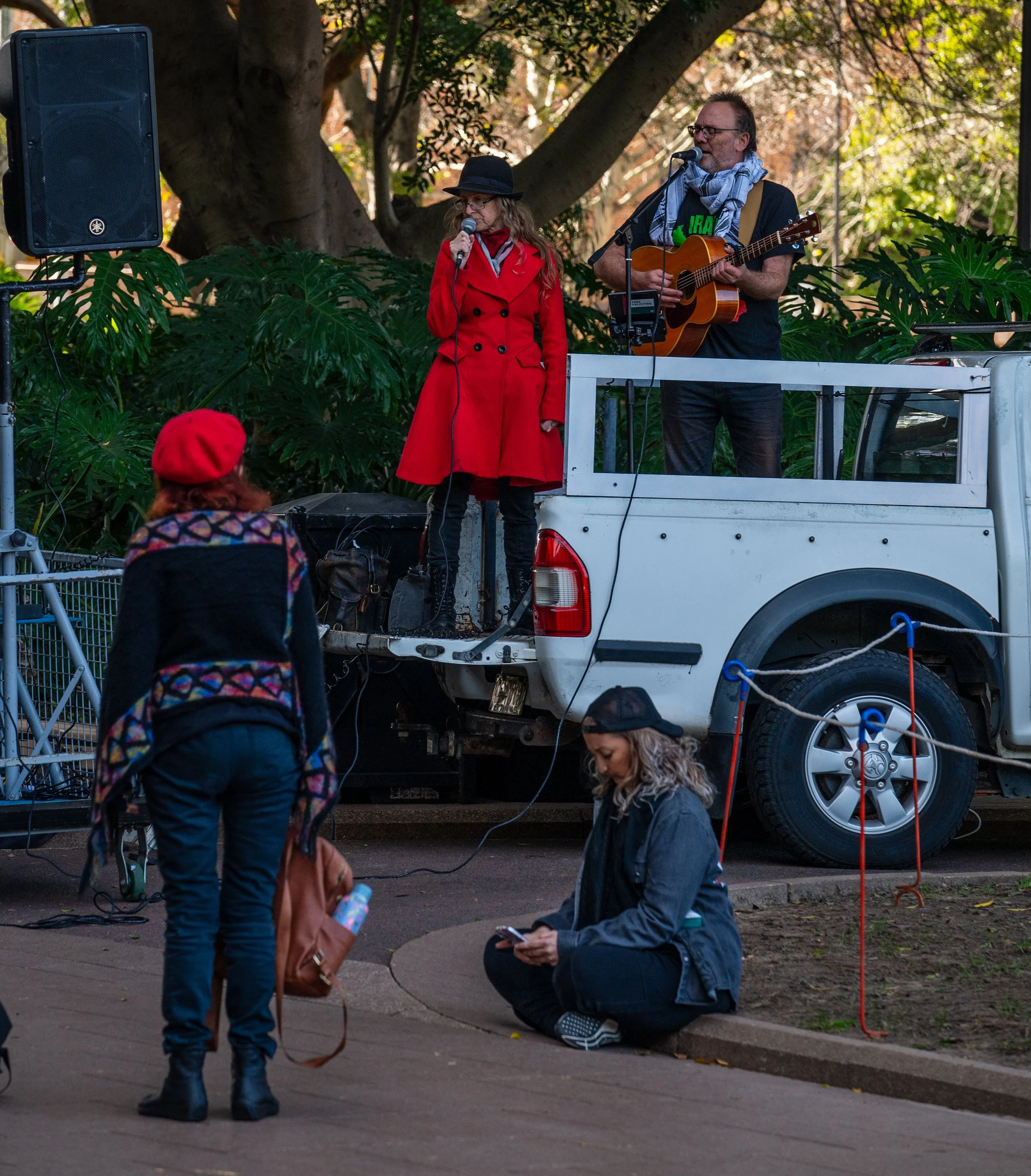 Two musicians perform on a small stage built on the back of a white pickup truck in an outdoor park. One woman sings into a microphone, dressed in a red coat and black hat, while the man plays an acoustic guitar. There are several people watching, including a woman sitting on the sidewalk and another woman wearing a red hat and colorful sweater standing nearby.