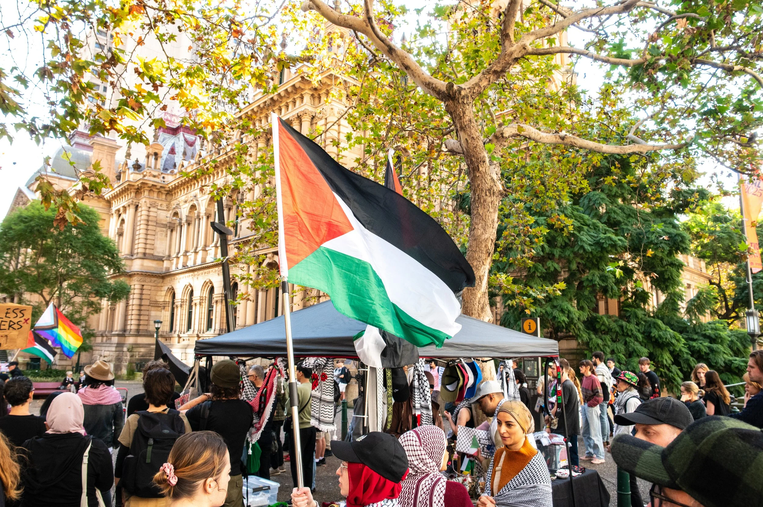 People gathered at an outdoor event with Palestinian flags and rainbow flags, surrounded by trees and historic buildings.