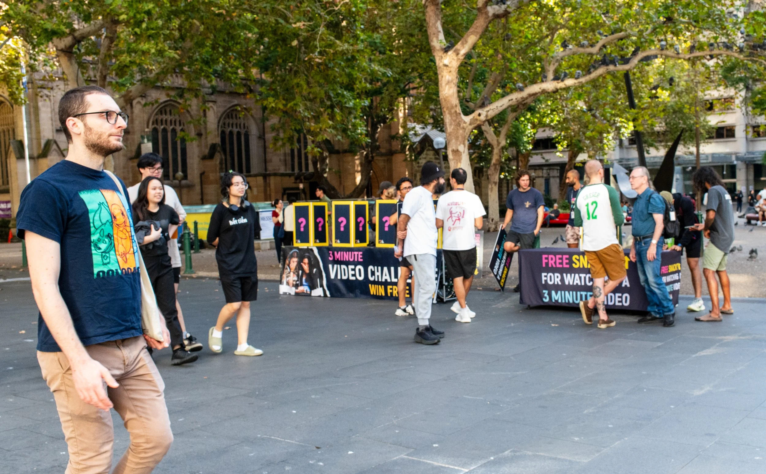People gathered in an outdoor city square participating in a promotional event for a 3-minute video challenge, with banners advertising free snacks for watching a 3-minute video, and some individuals engaging in conversations.