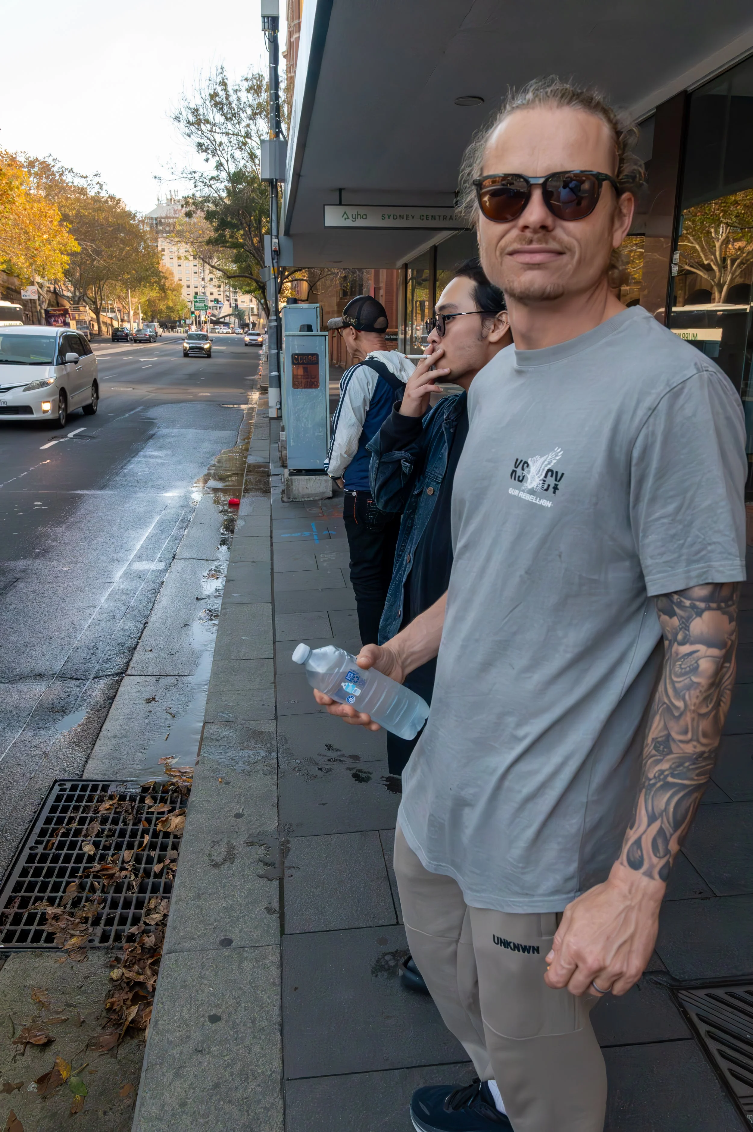 Group of people standing on a city sidewalk next to a street with passing cars, some with sunglasses, one person holding a water bottle, and trees with autumn leaves in the background.