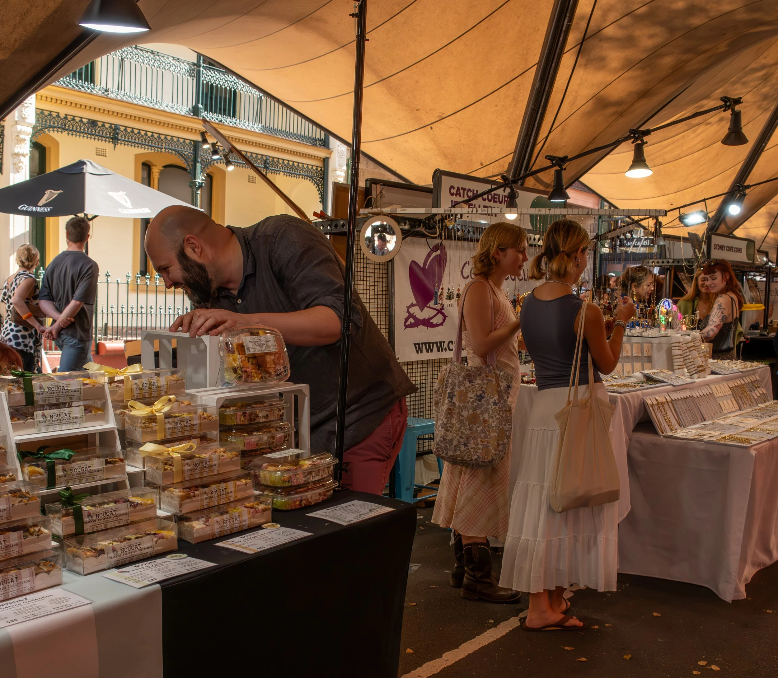 A street market stall selling baked goods, with customers browsing and purchasing, under a large tan canopy during daytime.