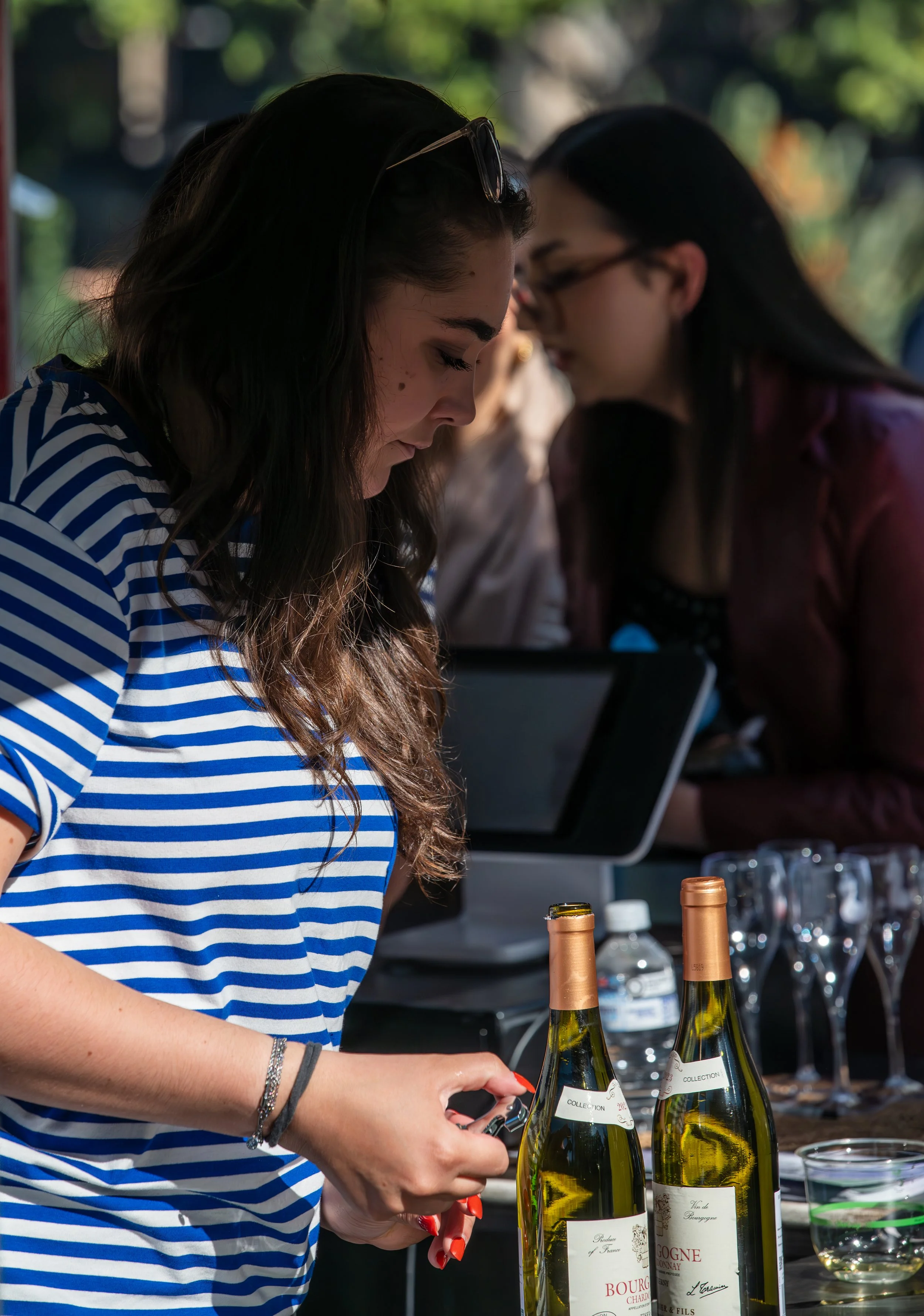 A woman with long brown hair wearing a blue and white striped shirt is opening a bottle of wine at a table, with two bottles of wine, glasses, and a water bottle in front of her. In the background, another woman with long black hair and glasses is looking at her tablet.