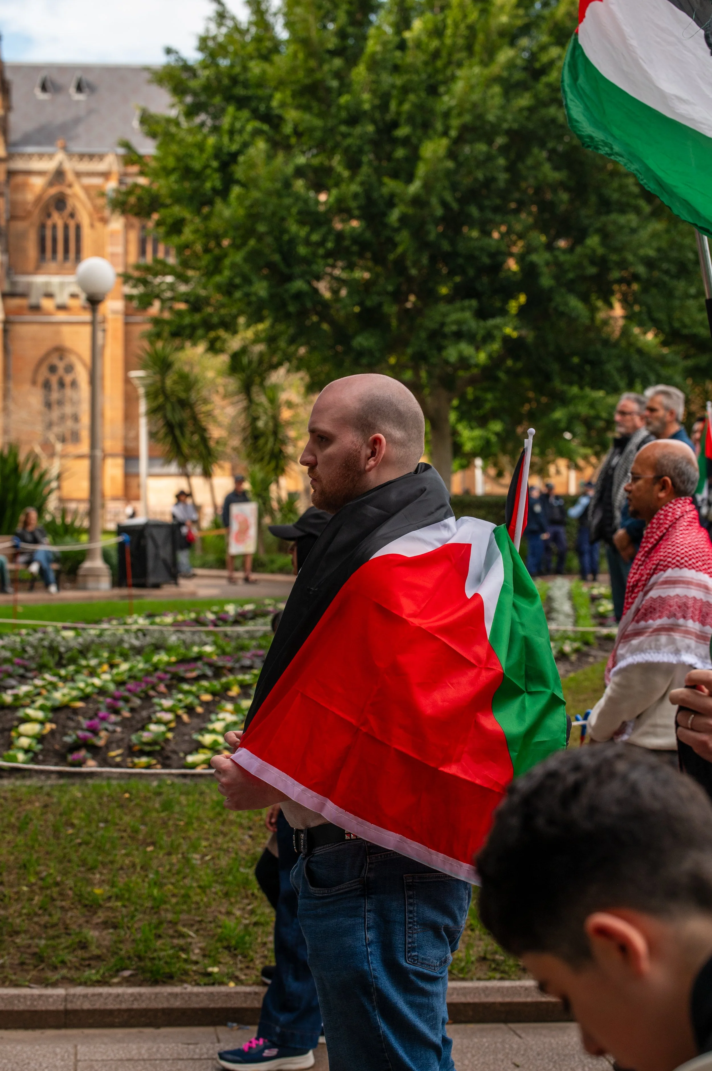 A man with a shaved head holding a flag that resembles the Palestinian flag, standing among a group of people in a park during a gathering or demonstration.