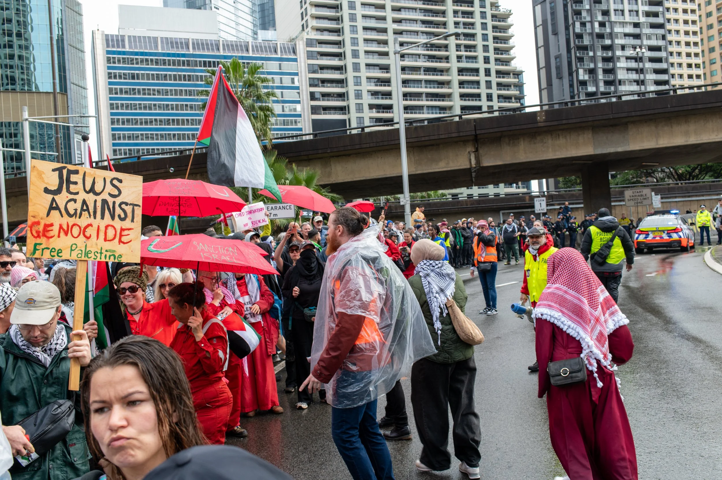 Protest on a city street with people holding signs and umbrellas, including a sign that reads 'Jews Against Genocide Free Palestine' and a Palestinian flag, with police and high-rise buildings in the background.
