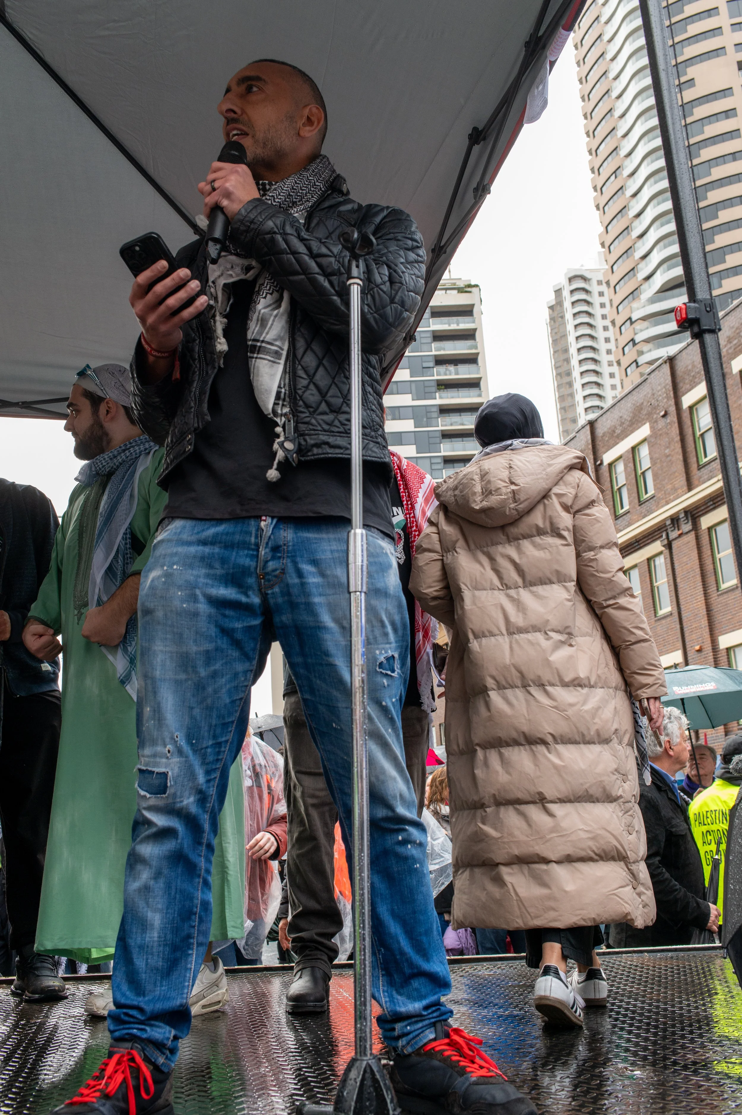 A man speaking into a microphone on a raised platform during a protest, holding a phone, with other protesters and tall buildings in the background.