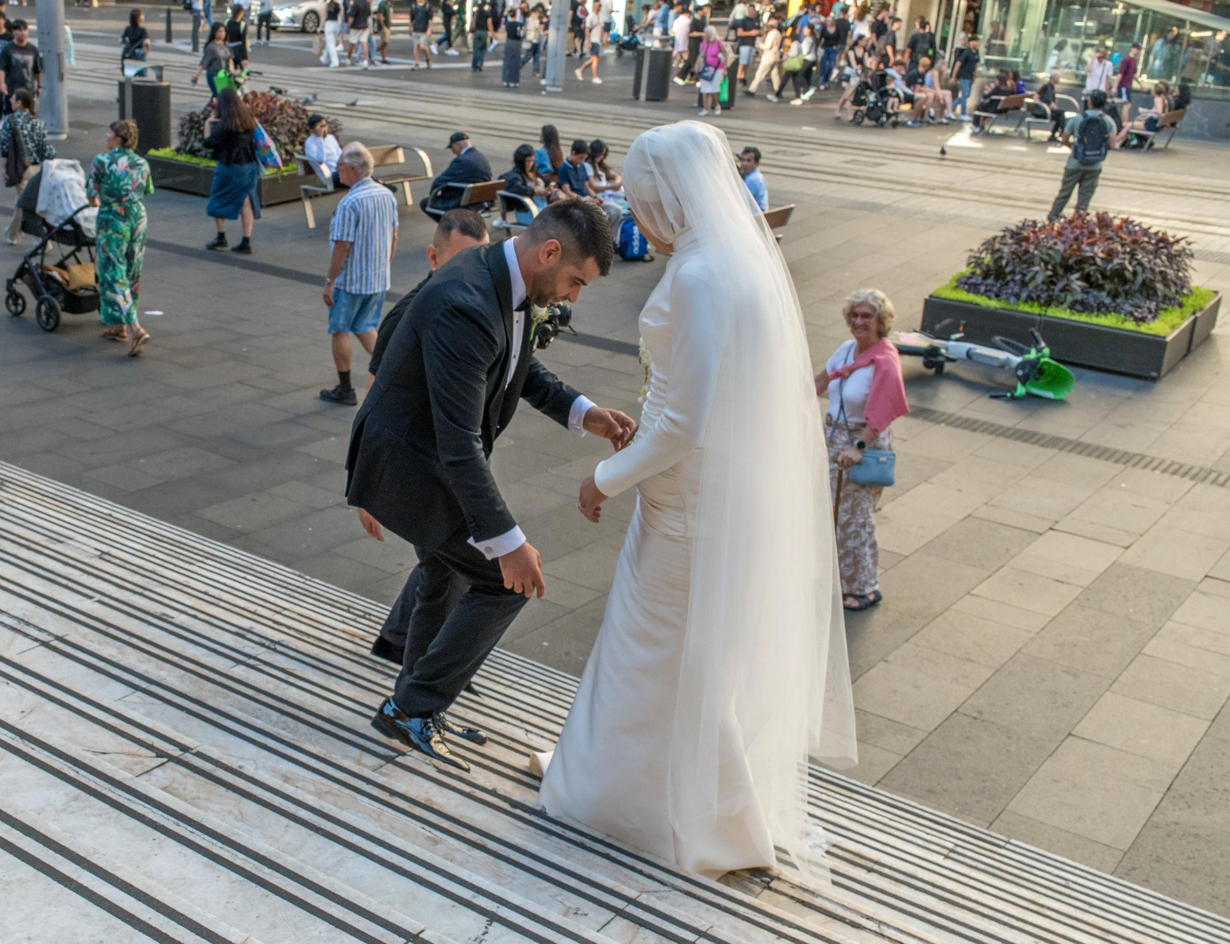 A man helping a woman in a white wedding dress down the steps in a busy city area filled with pedestrians.