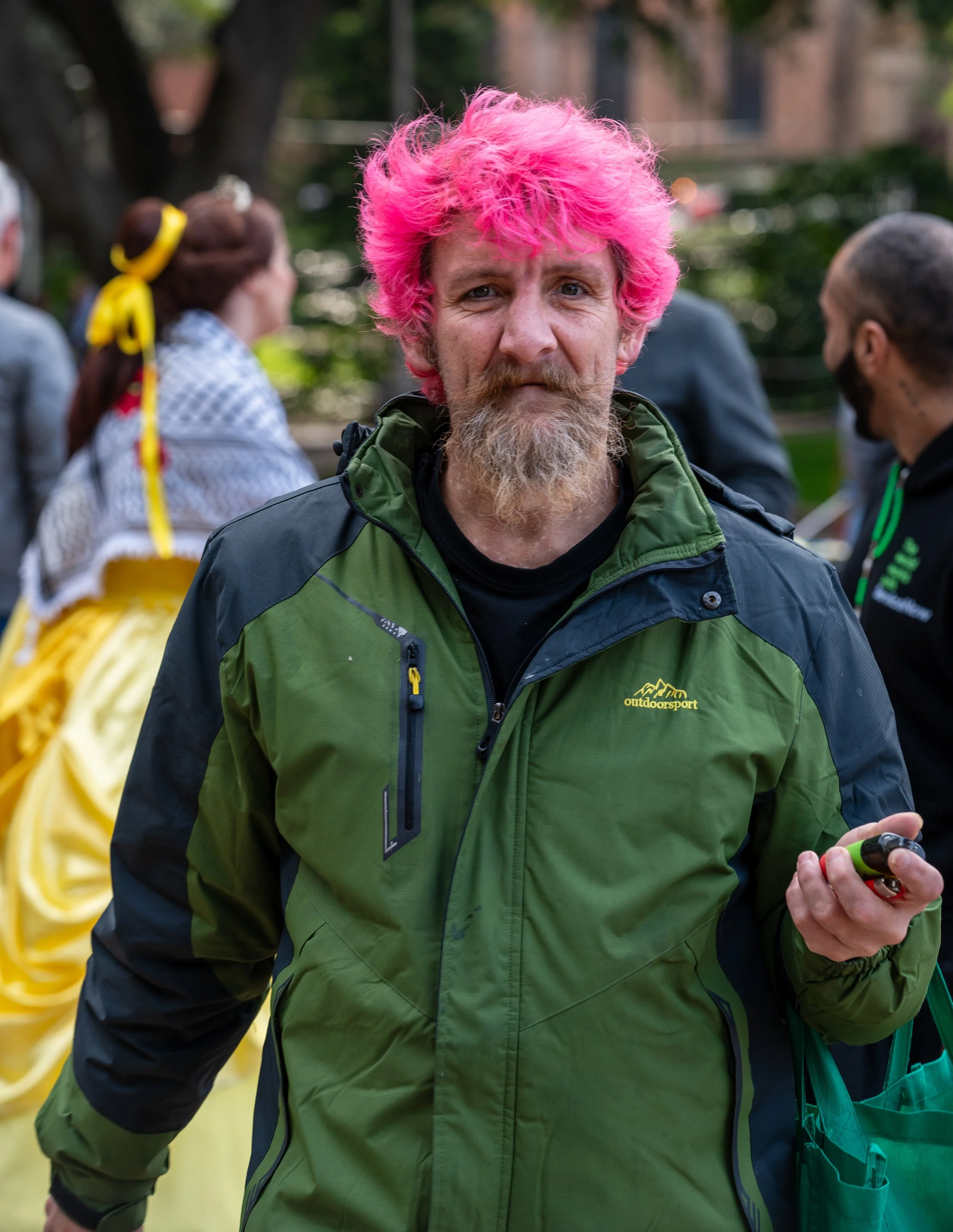 Man with pink hair and a beard wearing a green outdoor jacket, standing outdoors with blurred people in the background.