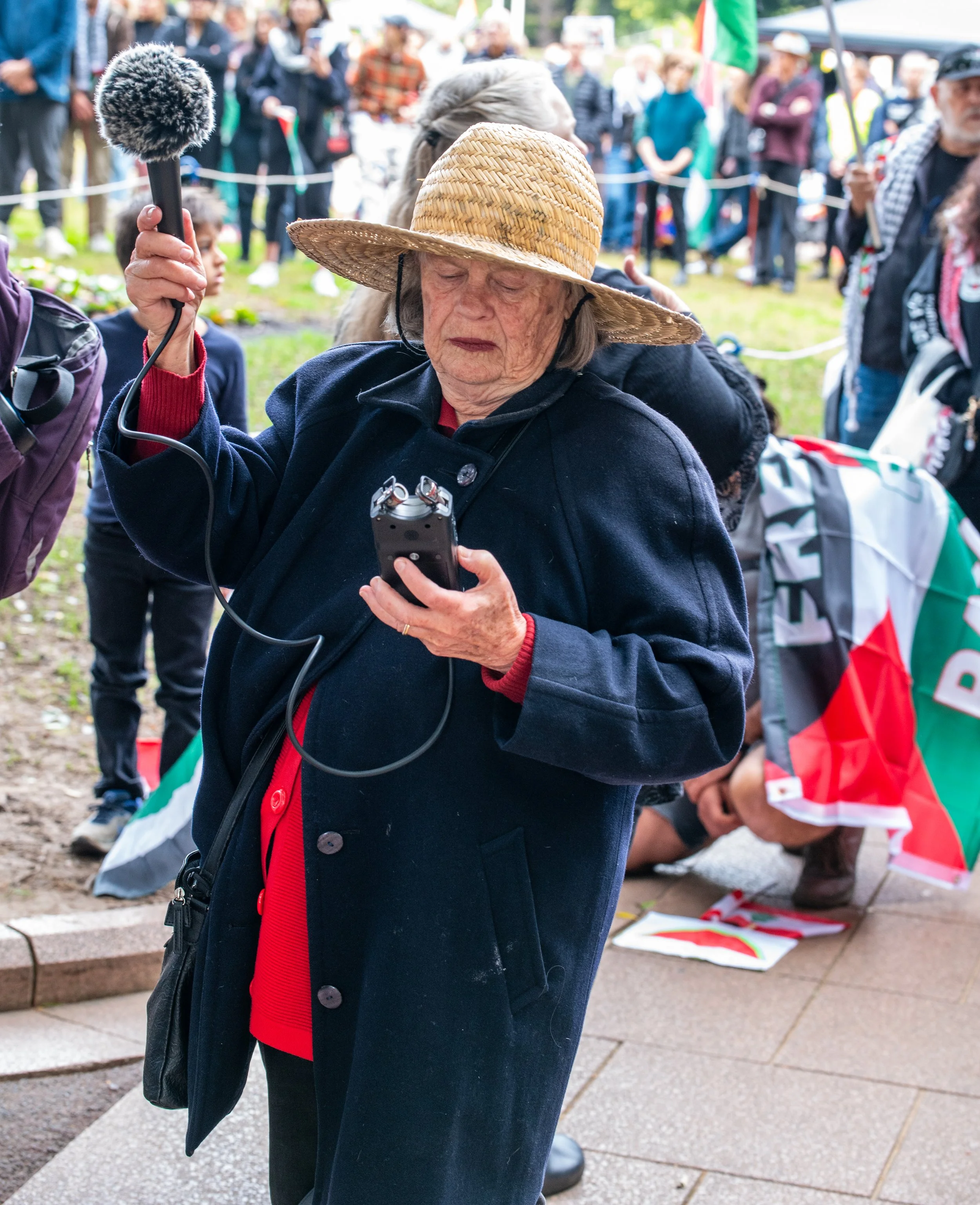 An elderly woman wearing a straw hat and dark coat at a protest or rally, holding a small electronic device connected to a microphone or recording device, with a crowd of people in the background.