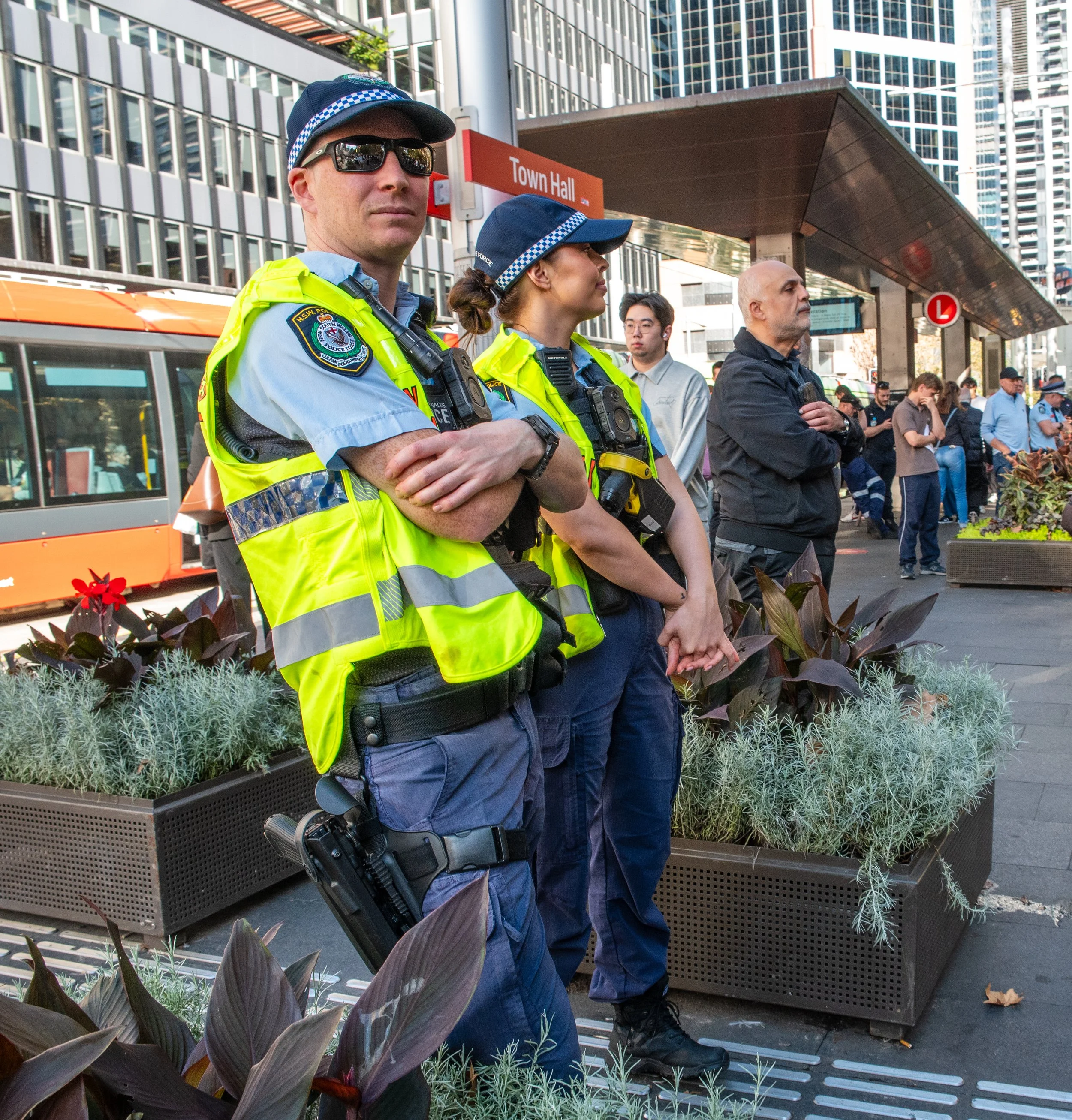 Two police officers in high-visibility vests and hats standing with arms crossed at a busy urban street corner, with multiple pedestrians and a tram in the background.