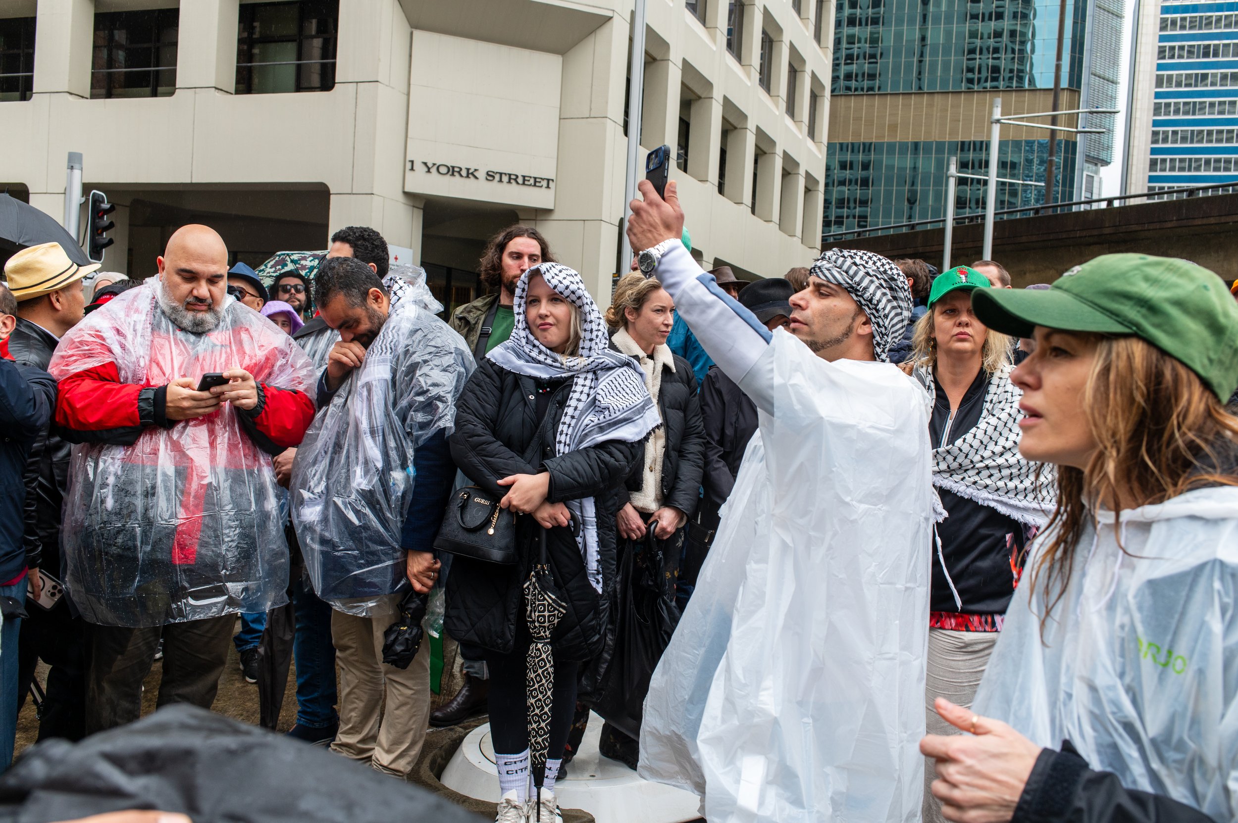 People gathered at a protest or rally in an urban setting, some wearing rain ponchos and headscarves, with buildings and a street sign reading '1 York Street' in the background.