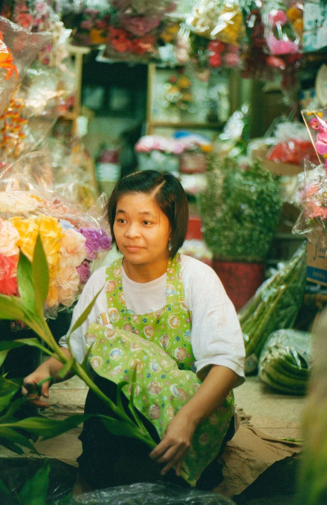 A woman with short dark hair and a light skin tone working in a flower shop, holding a bouquet of colorful flowers, surrounded by various floral arrangements and supplies.