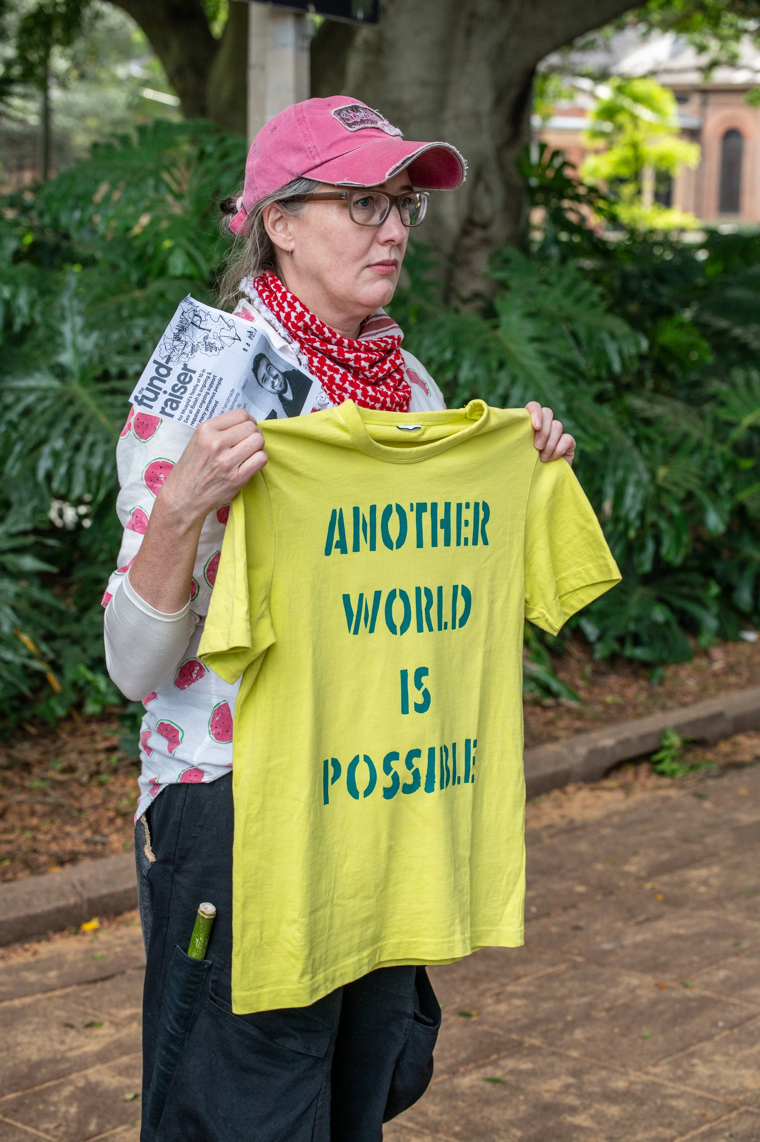 A woman wearing glasses, a pink cap, and a red and white scarf holds a bright yellow T-shirt with the words 'ANOTHER WORLD IS POSSIBLE' printed on it. She also holds a pamphlet or flyer in her left hand. She stands outdoors in front of green foliage and trees.