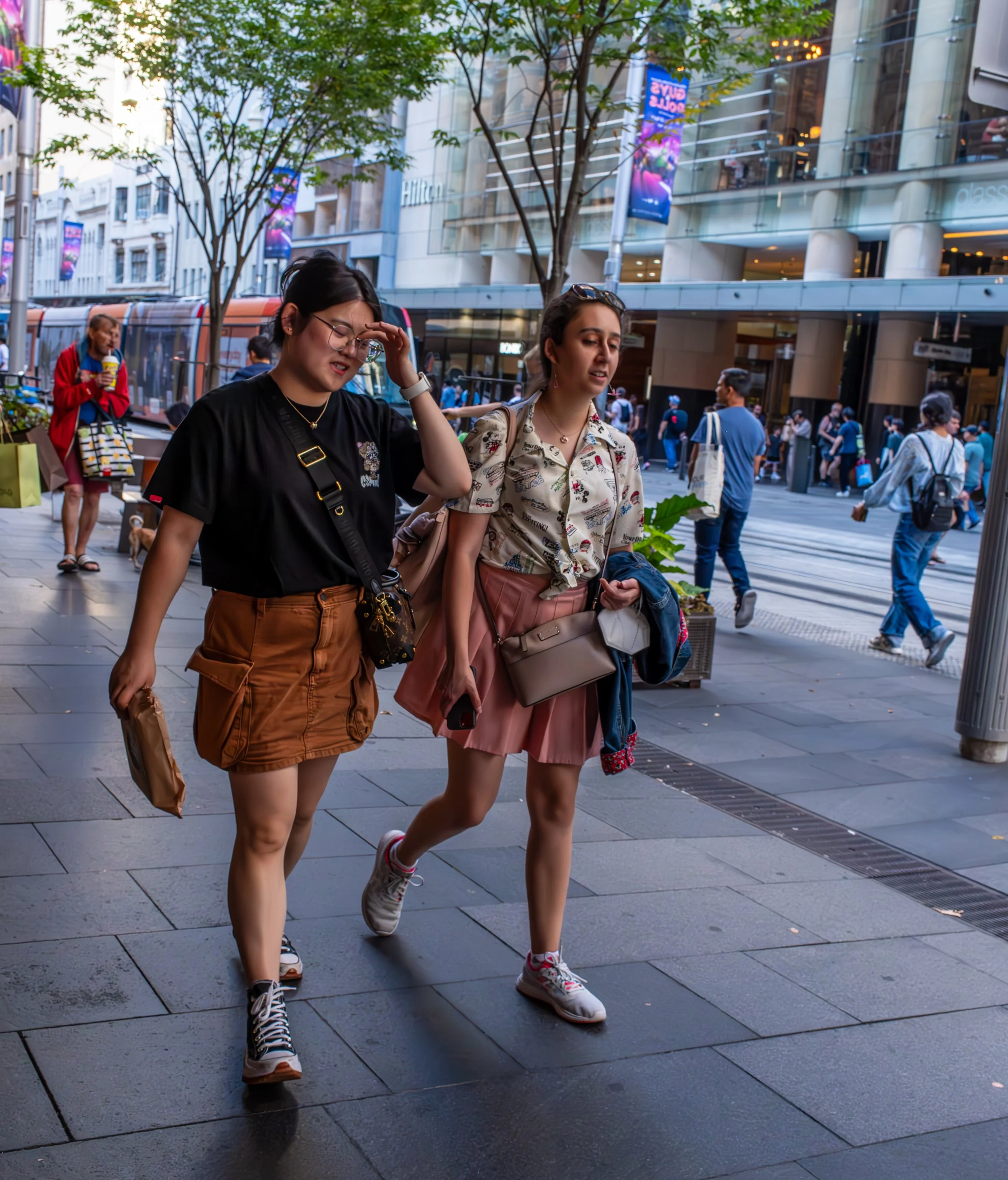 Two young women walking on an urban street, one adjusting her glasses, the other holding a phone and a jacket, with a busy city scene in the background.
