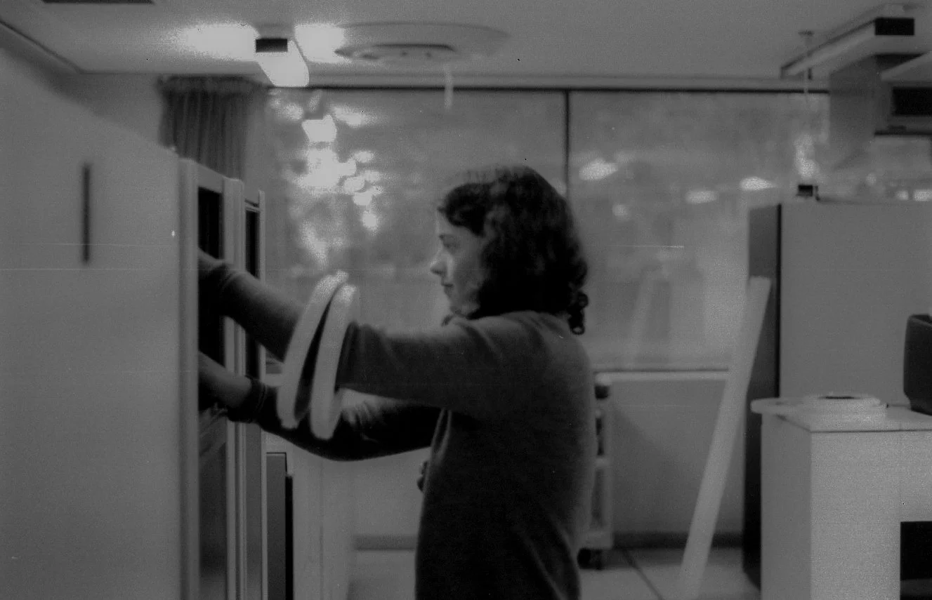 A woman with dark, curly hair reaching into a mailbox or deposit box in an office setting, with a glass wall behind her reflecting ceiling lights.