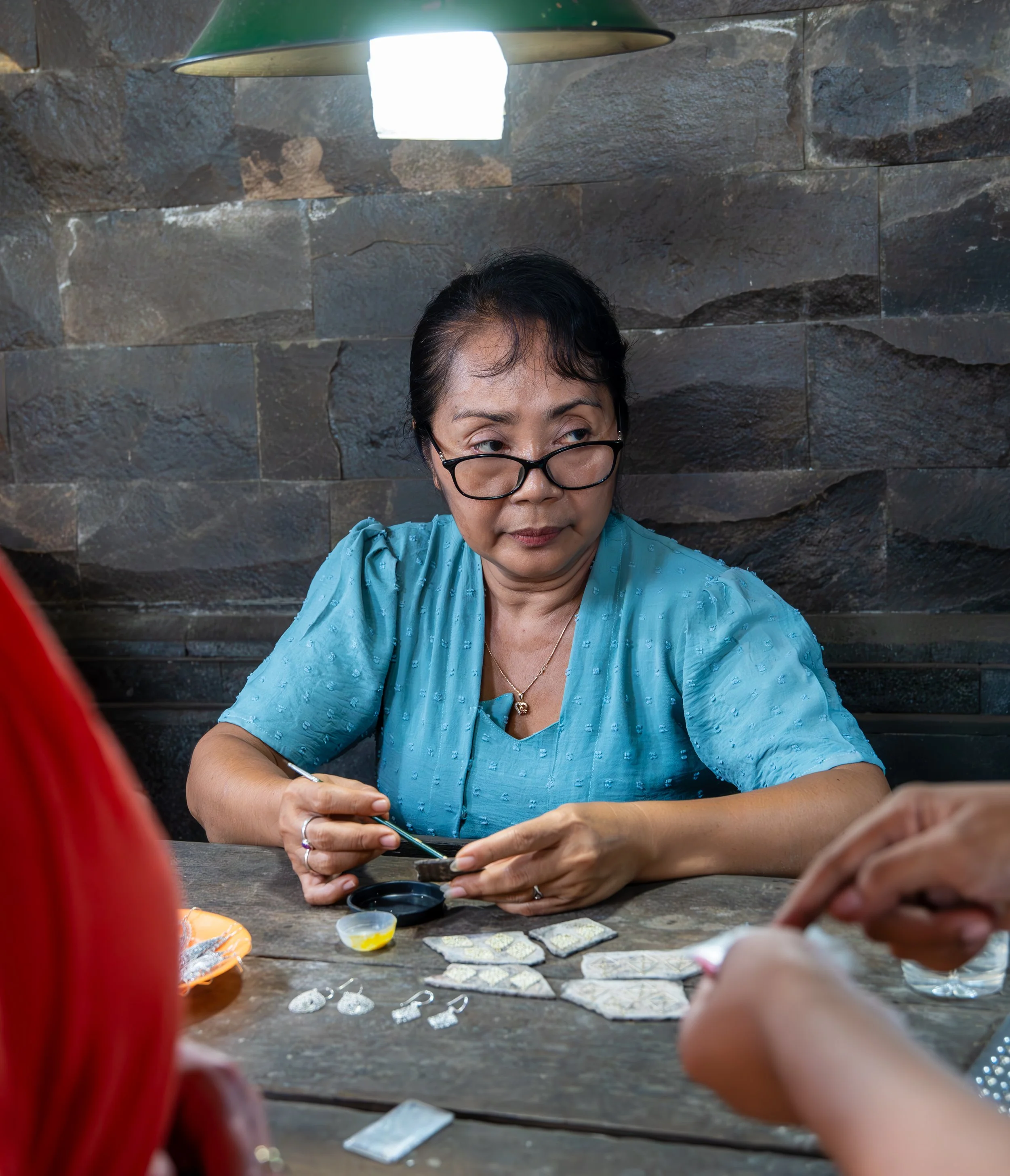 A woman with glasses and a blue shirt sitting at a table, making jewelry with beads and tools, with a stone wall background.