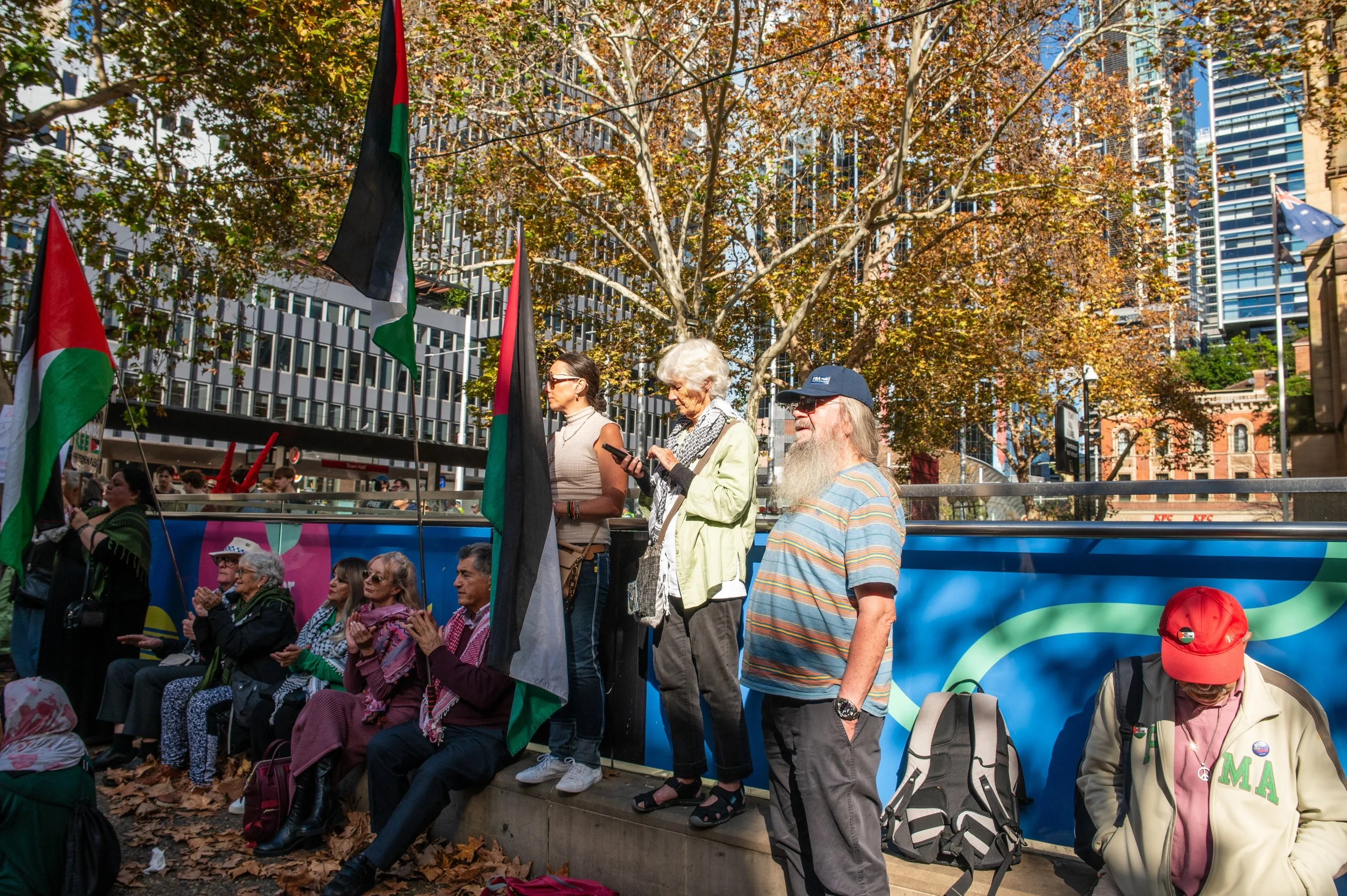 A group of people gathered outdoors at a rally or protest, some standing and others sitting, with various flags, including Palestinian flags, and trees with autumn leaves, city buildings in the background, and it appears to be daytime.