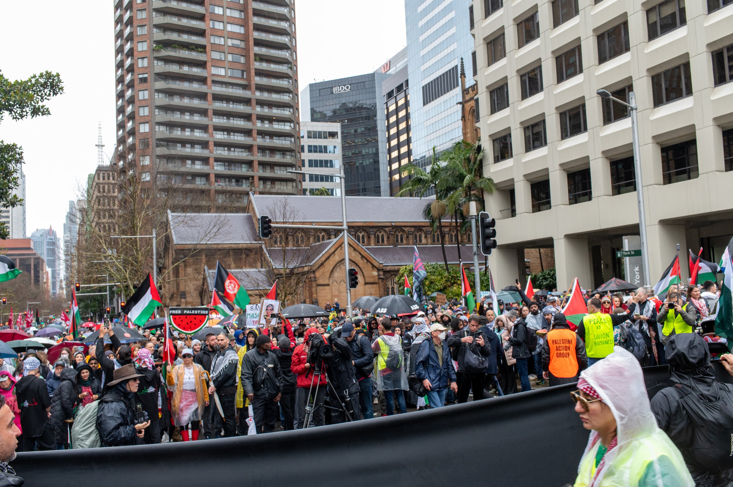 Protest march in city with people holding Palestinian flags, banners, and umbrellas, some wearing raincoats, urban high-rise buildings in the background.