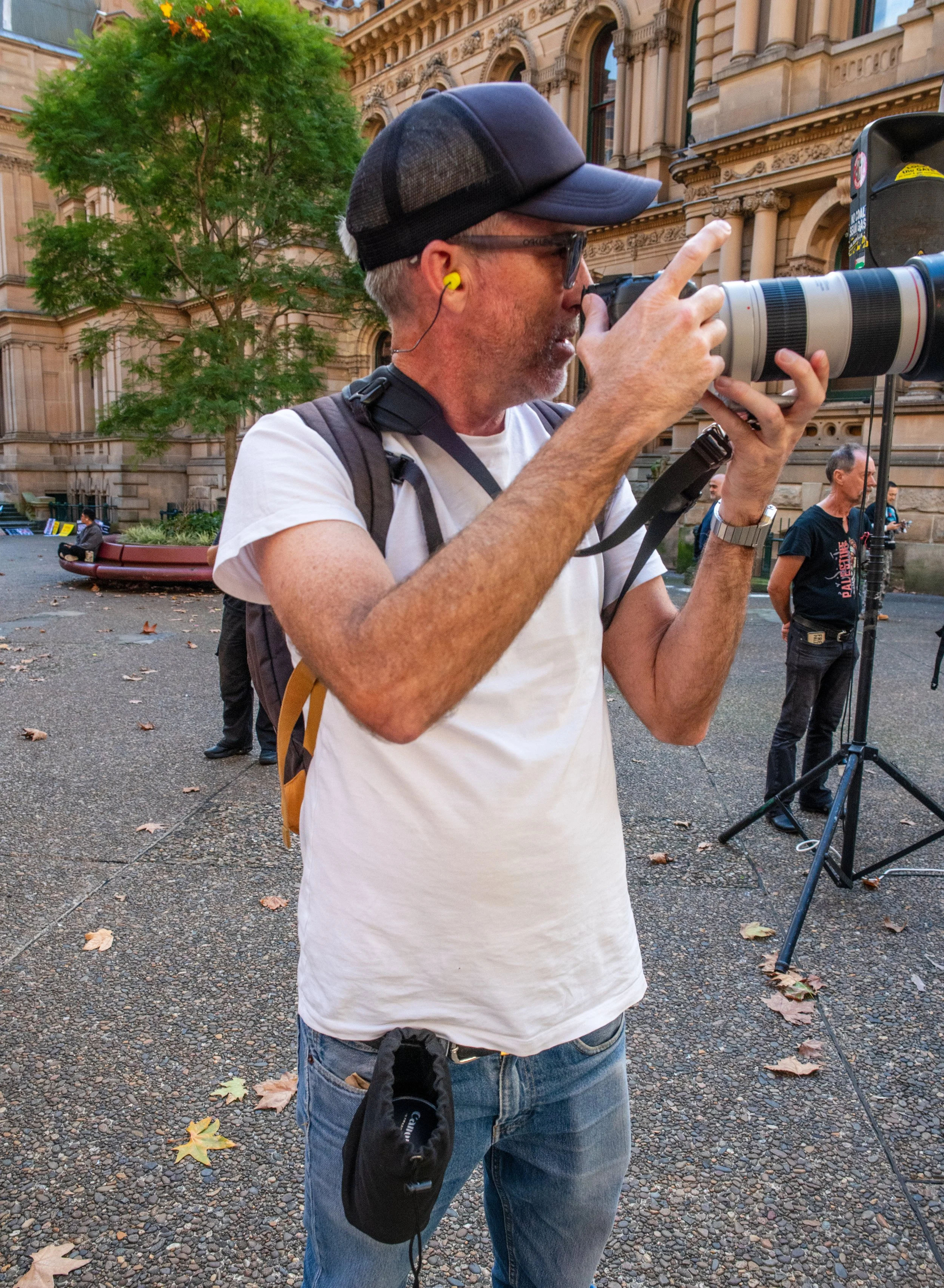 A man wearing a baseball cap, sunglasses, a white t-shirt, and yellow ear protection is taking a photograph with a large telephoto lens camera outdoors in front of a historic building. There are other people in the background, some standing and some sitting, on the sidewalk, and a tree with green leaves is visible.