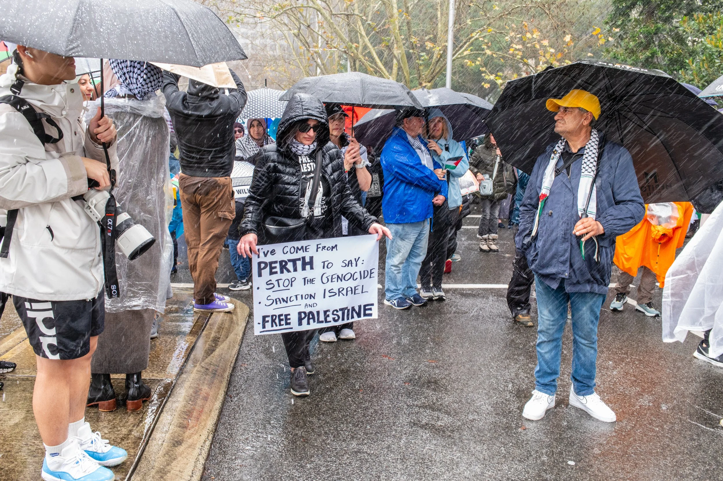 People protesting in rain with umbrellas, one woman holding a sign that reads 'I've come from Perth to say: Stop the genocide, sanction Israel, and free Palestine'