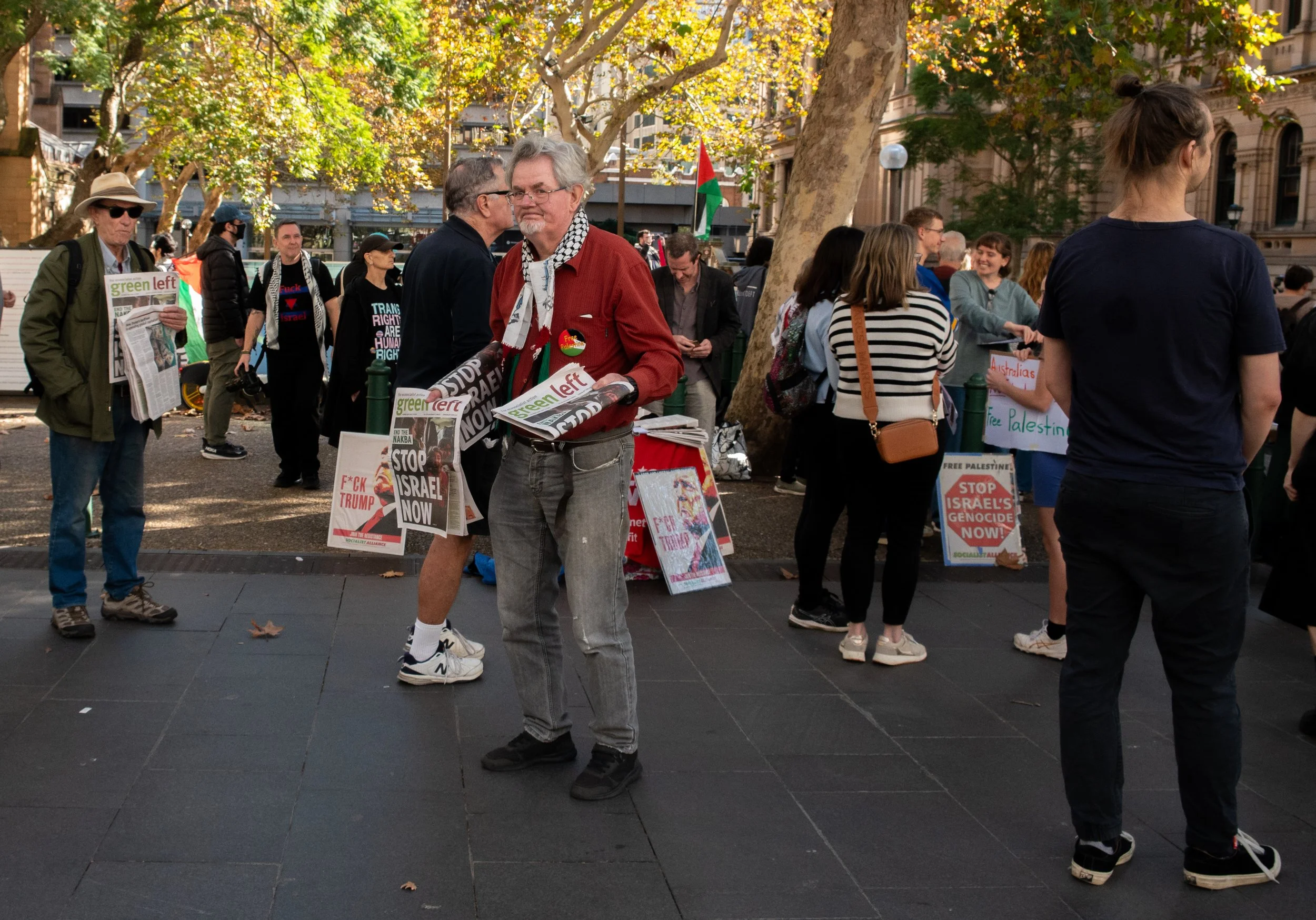 Protesters gathered in a park holding signs with messages calling to stop Israeli actions, supporting Palestine, and opposing Trump, with some signs reading "Stop Israel Now" and "Free Palestine". Some protesters are talking, reading, or holding signs, with trees and buildings in the background.