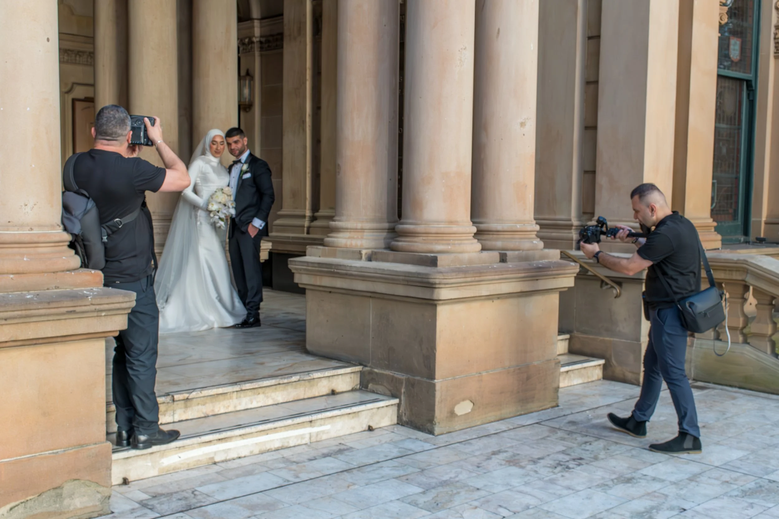 A bride and groom standing on steps outside a building with large columns, taking a photo while two photographers capture the moment.