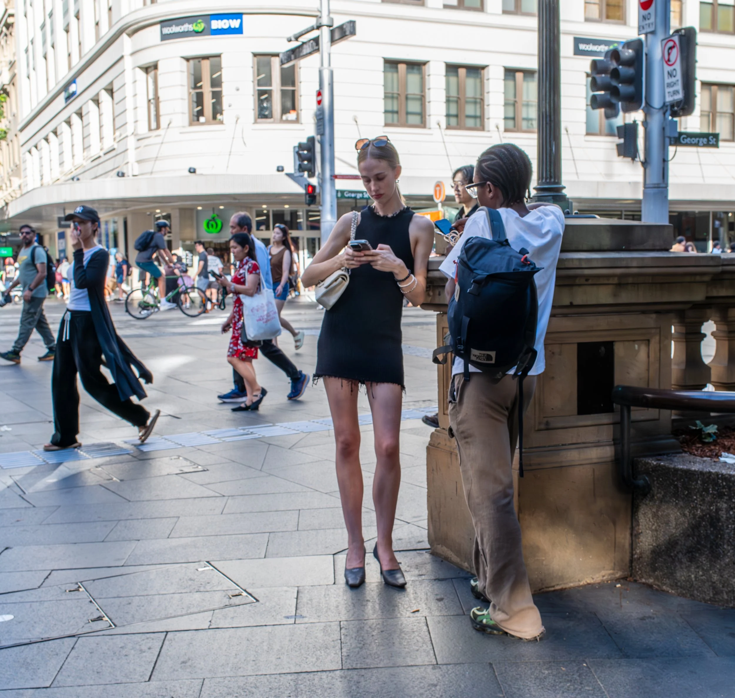 Two women are standing on a city street, one in a black dress looking at her phone, and the other in a white shirt with a backpack, talking to her. Pedestrians walk in the background, and traffic lights and street signs are visible.