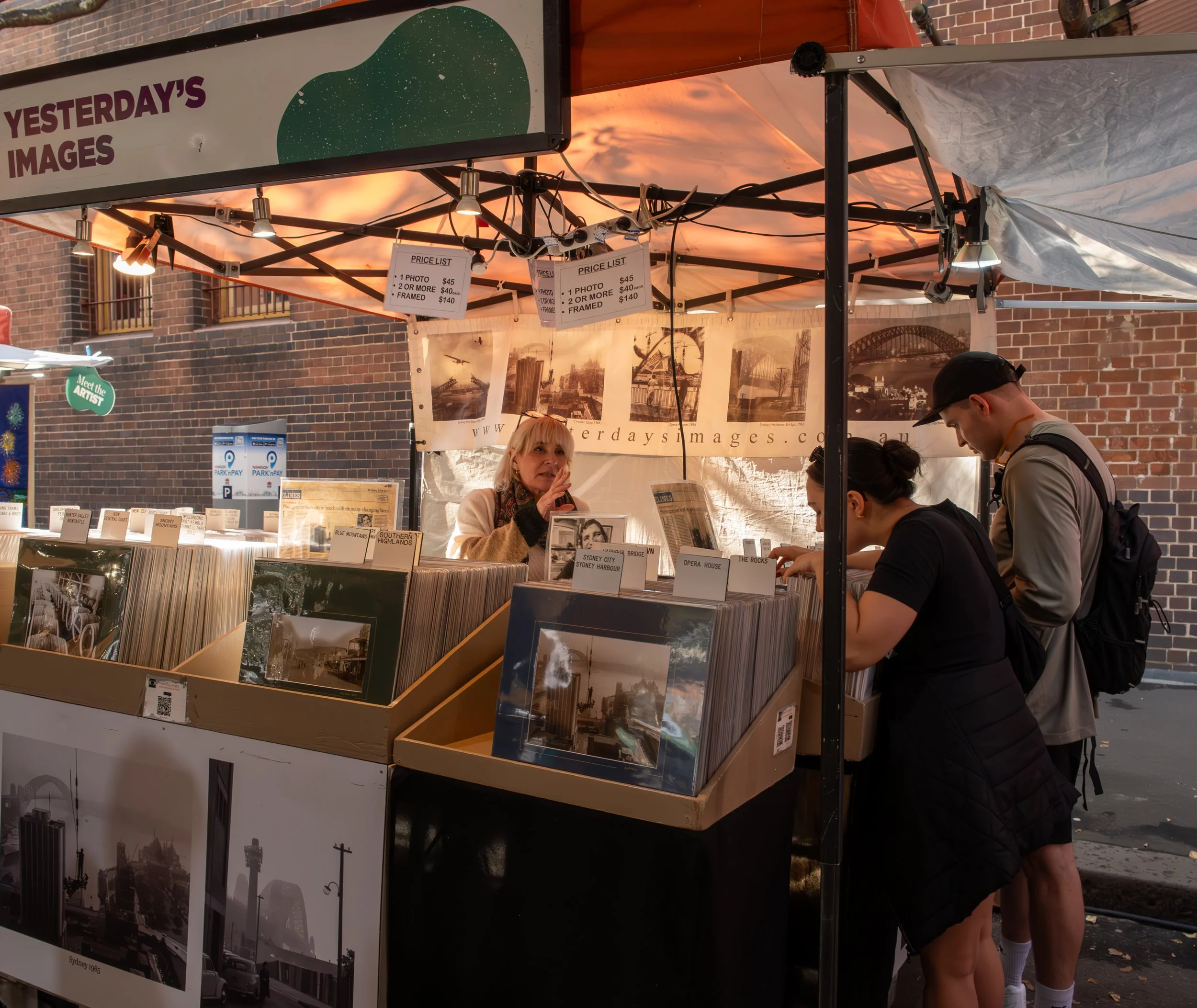 A street market booth selling black and white photographs, with two customers browsing and a vendor behind the counter. The booth has a sign reading 'YESTERDAY'S IMAGES' and displays various framed photos and photo prints.
