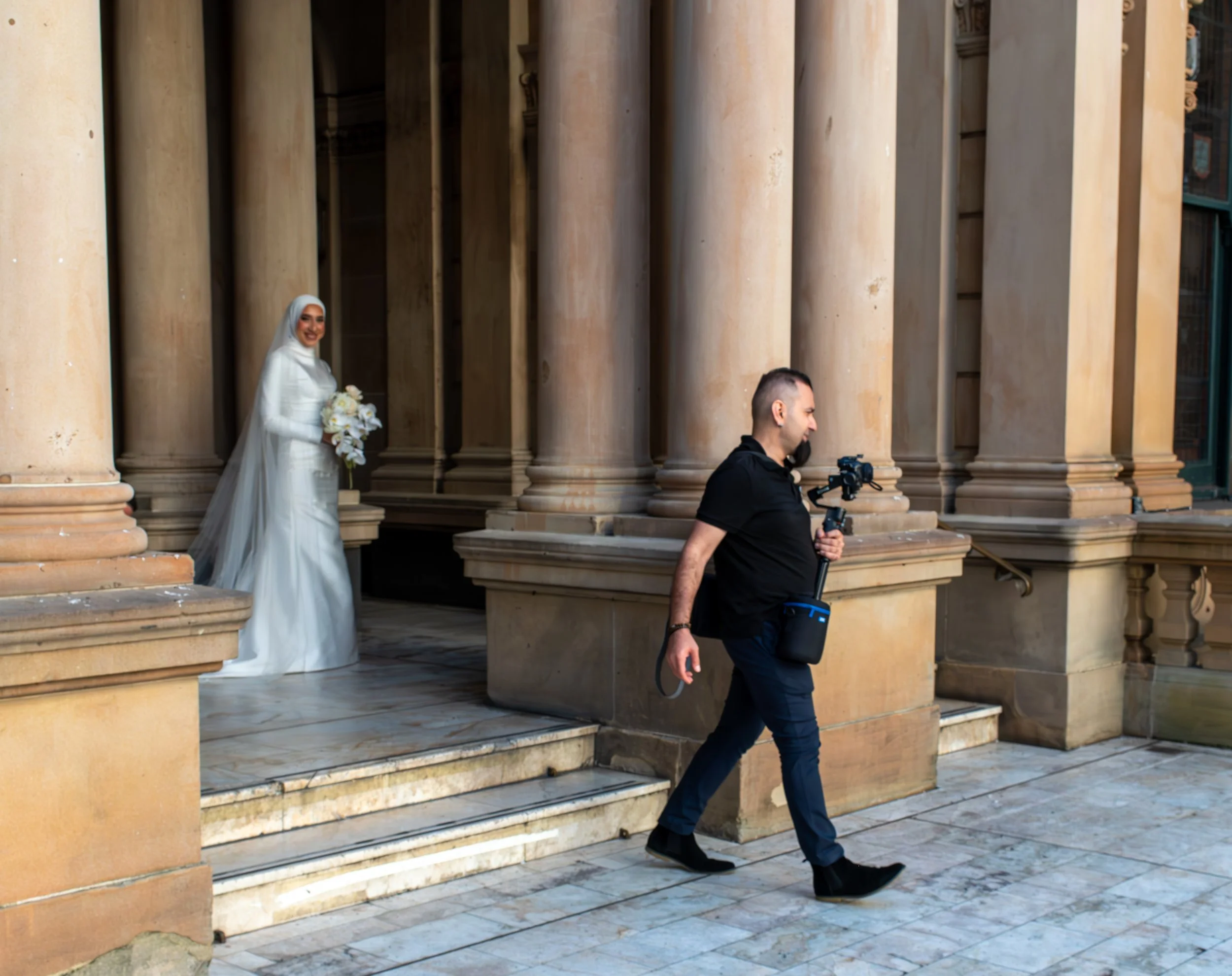 A bride in a white wedding dress and hijab holding a bouquet, smiling, as a cameraman walks past on a stone building with large columns.
