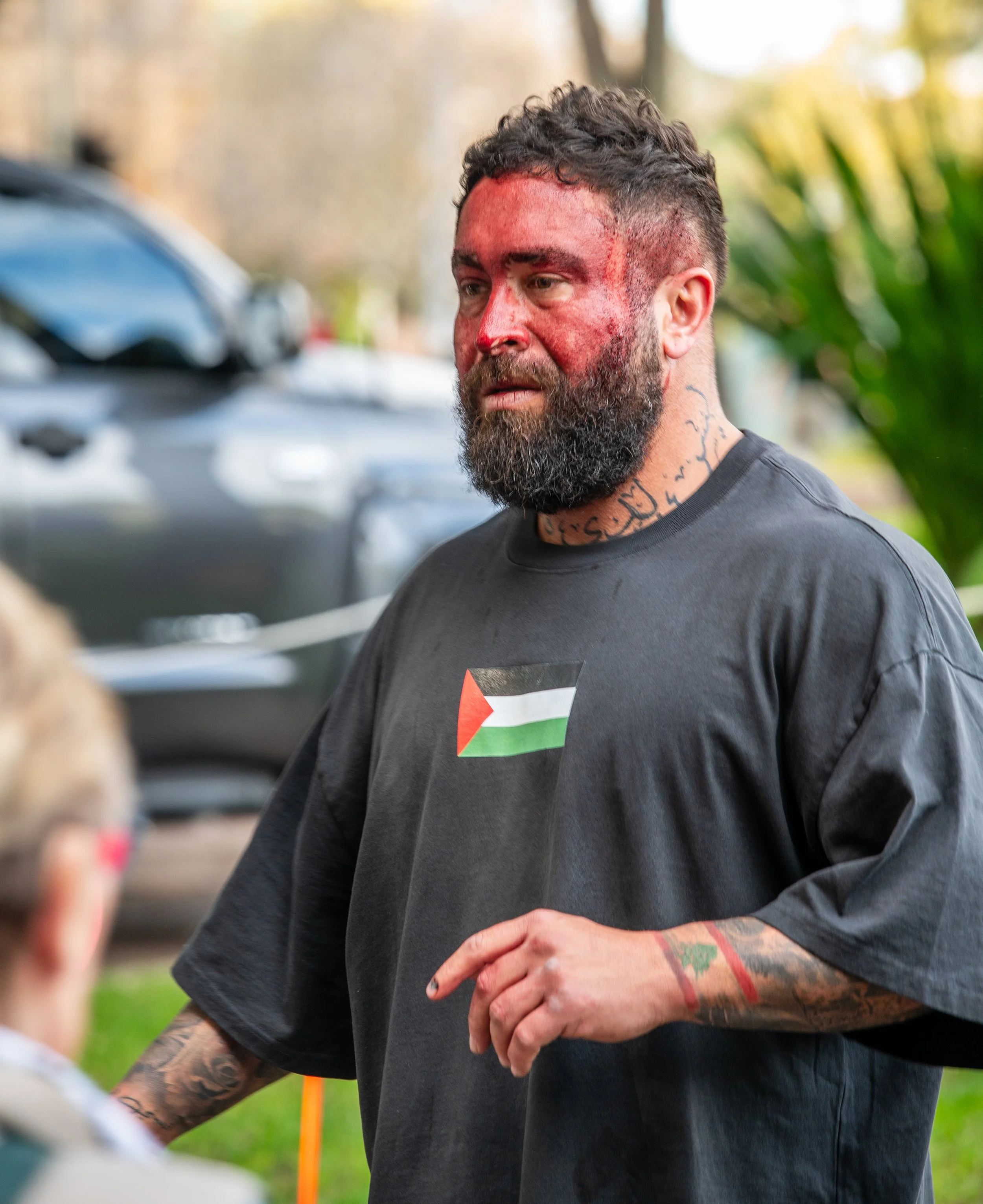 A man with a beard, tattoos, and a red face is talking outdoors, wearing a black shirt with a Palestinian flag on it.