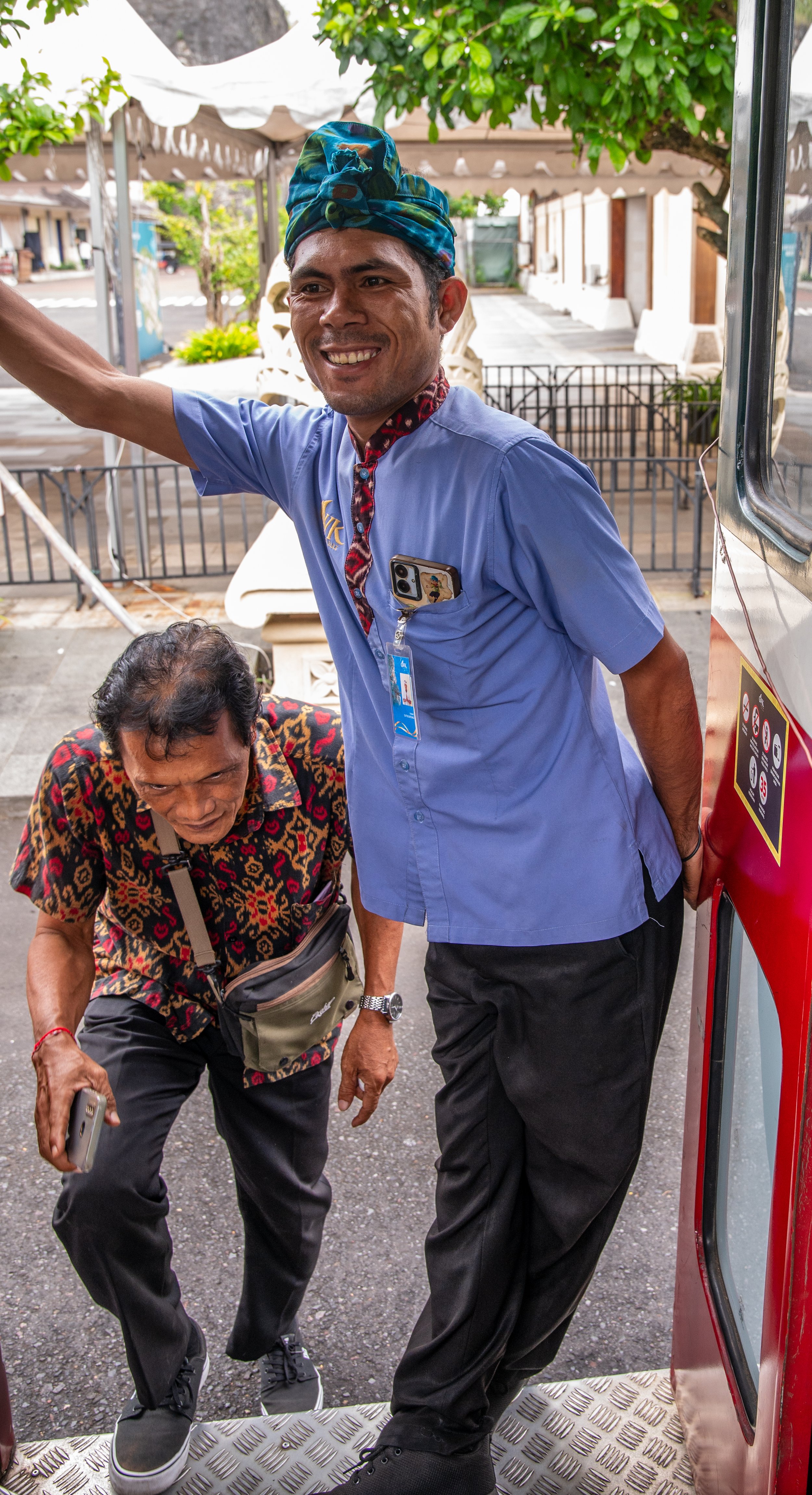 A smiling man in a blue shirt with a headwrap stands next to an older person with gray hair, wearing a patterned shirt and holding a phone, in an outdoor setting with tents and greenery.