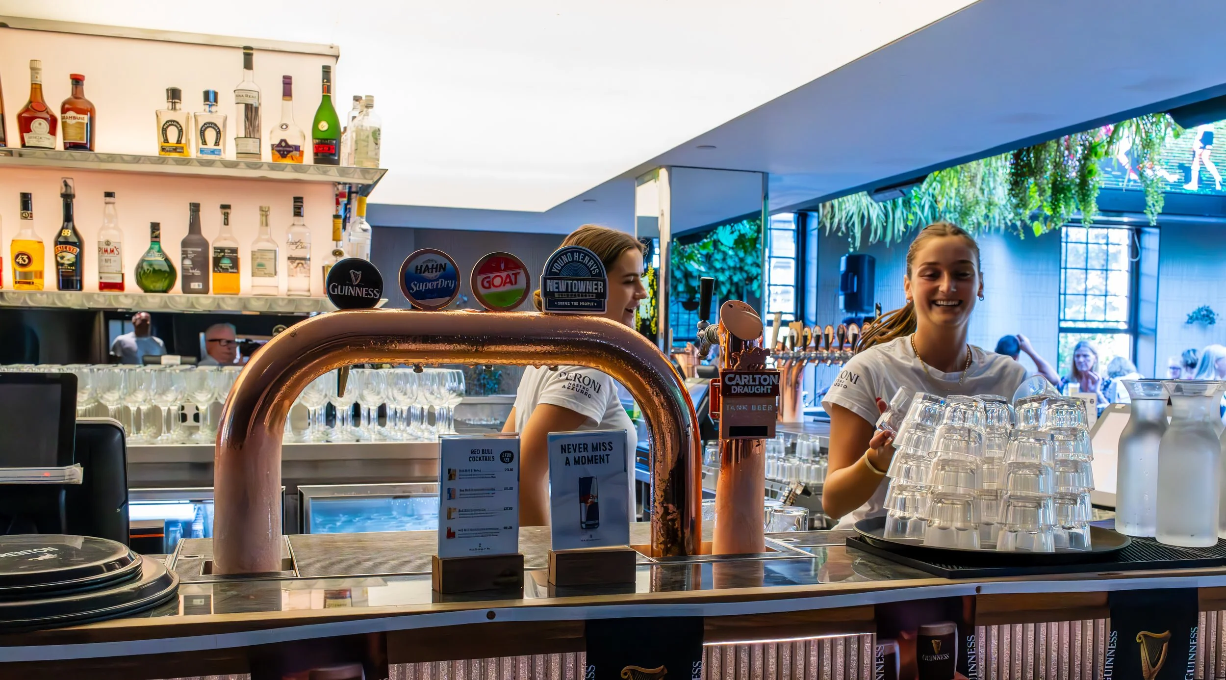 Bar with two smiling female bartenders behind the counter, shelves with liquor bottles, and multiple clean glasses on the counter, in a modern, well-lit setting with patrons in the background.