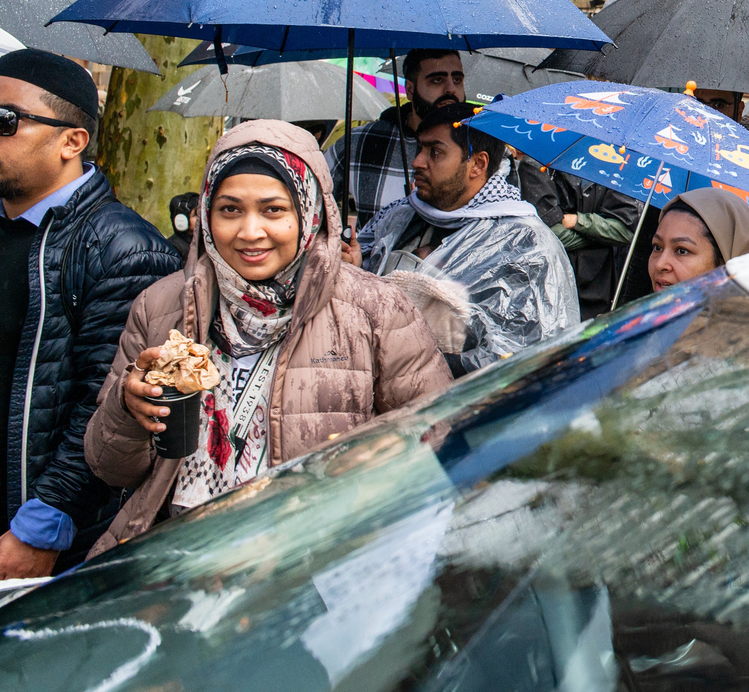 A diverse group of people gathered outdoors in the rain with umbrellas, some wearing rain ponchos, and one woman in the front holding a cup with a lid and crumpled paper, smiling at the camera.