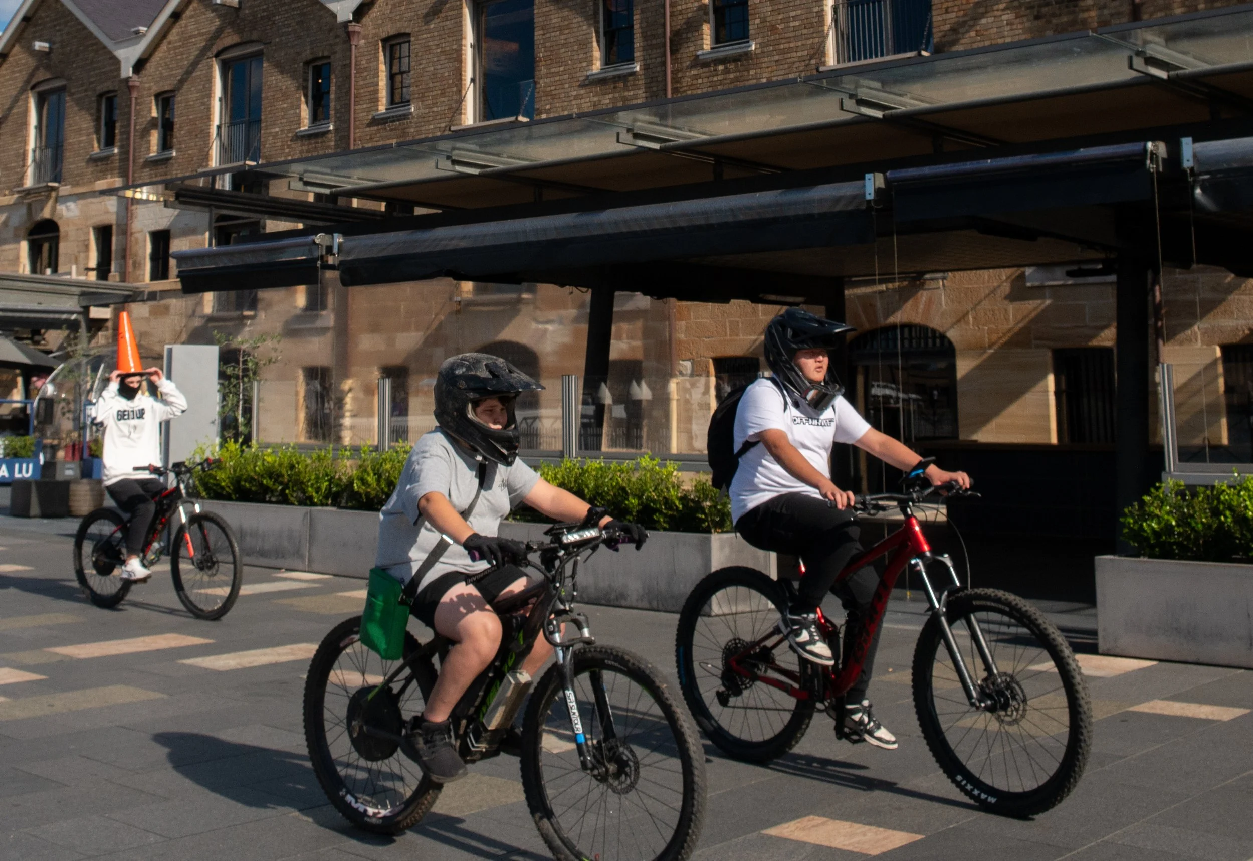 Three children riding bicycles on city street during daytime, with a person wearing an orange cone hat in the background.