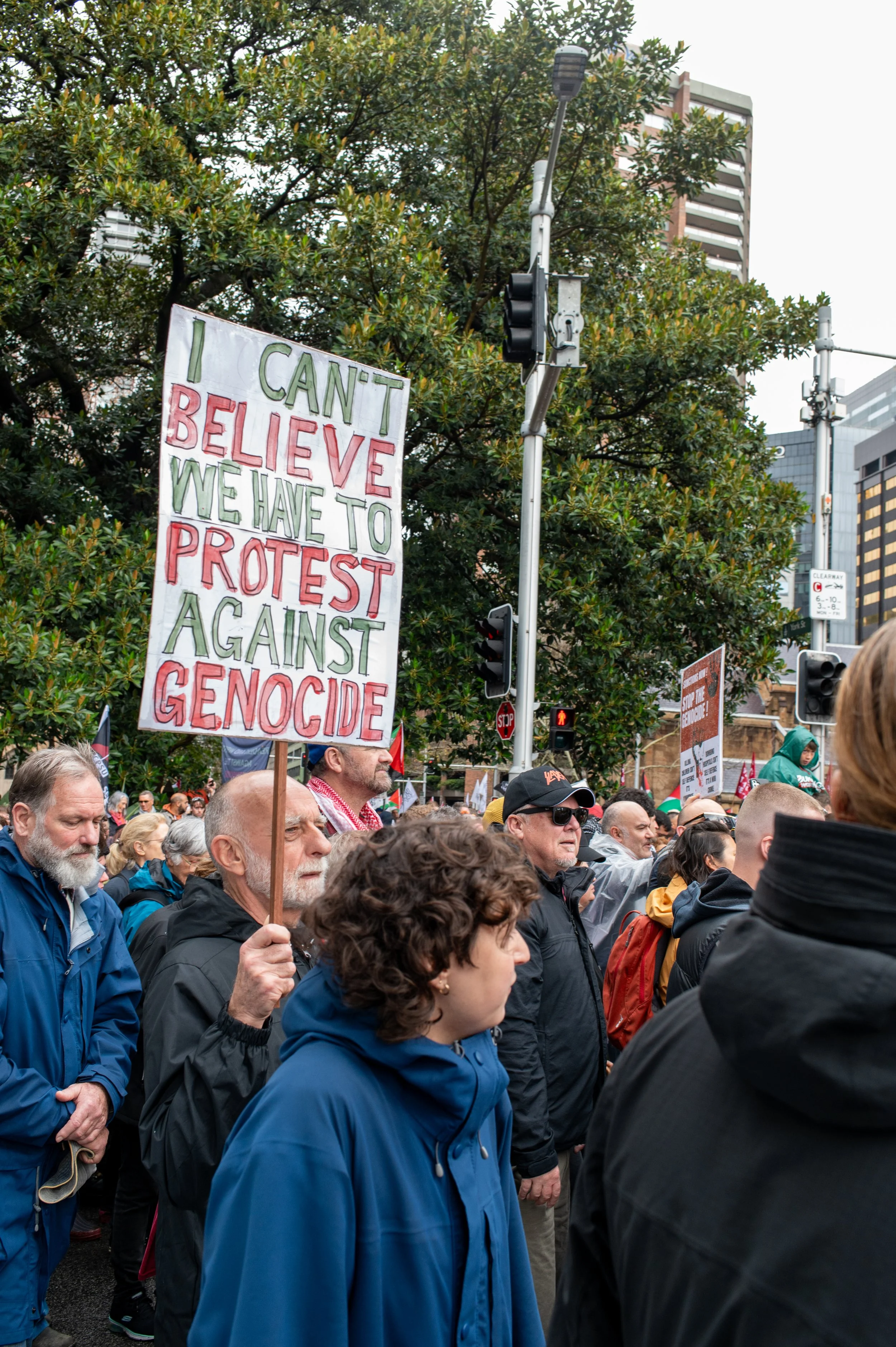 Protesters gathering outdoors, holding signs including one that reads 'I CAN'T BELIEVE WE HAVE TO PROTEST AGAINST GENOCIDE', in an urban setting with traffic lights, buildings, and trees.
