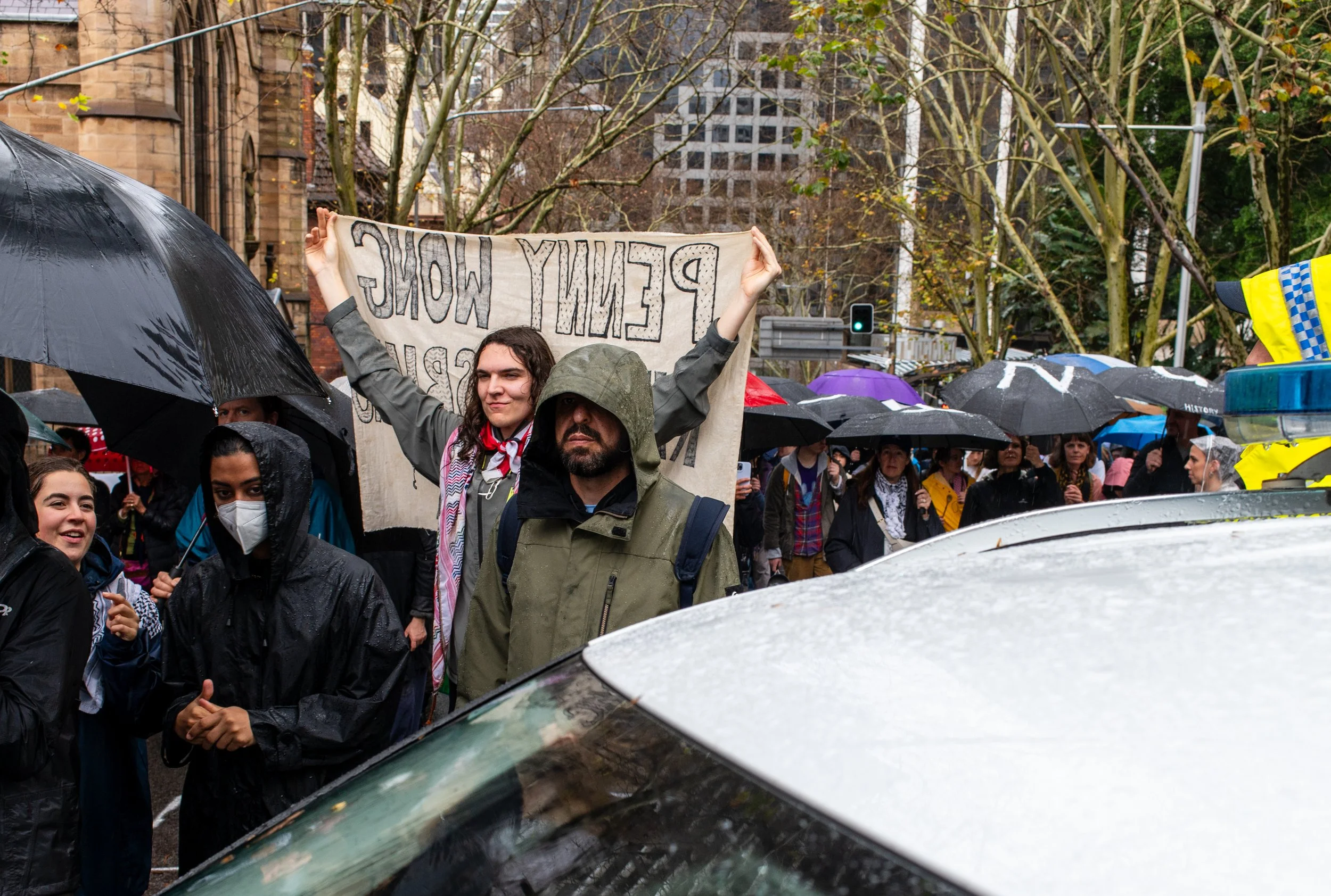 A group of protesters holding umbrellas in rainy weather, with a person in a gray hooded jacket lifting a large sign that reads 'PEMIFY WOLF' and others standing behind. The scene is in an urban area with trees and tall buildings in the background.