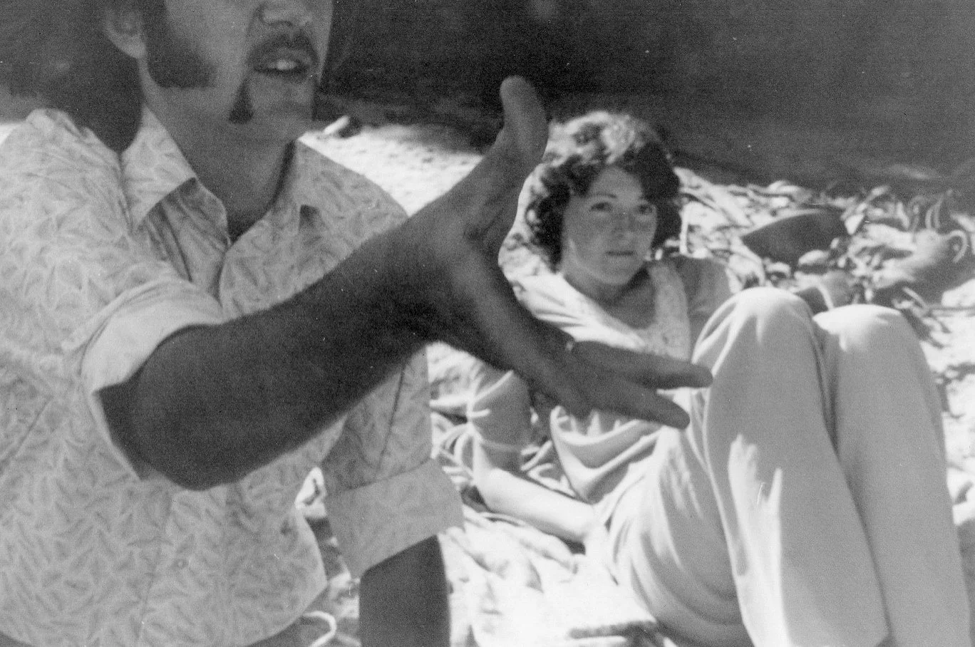 A black and white photo of a man with a beard and a woman with short, wavy hair on a rocky beach, with the man gesturing towards the camera and the woman sitting by the shore.